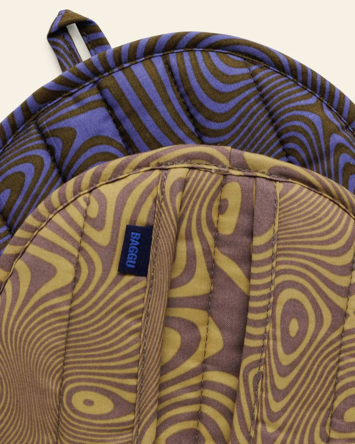 The BAGGU Pot Holder Set of 2 features quilted organic cotton mitts in wavy, psychedelic patterns—one purple and brown, one yellow and brown—both with looped handles and a blue BAGGU tag.