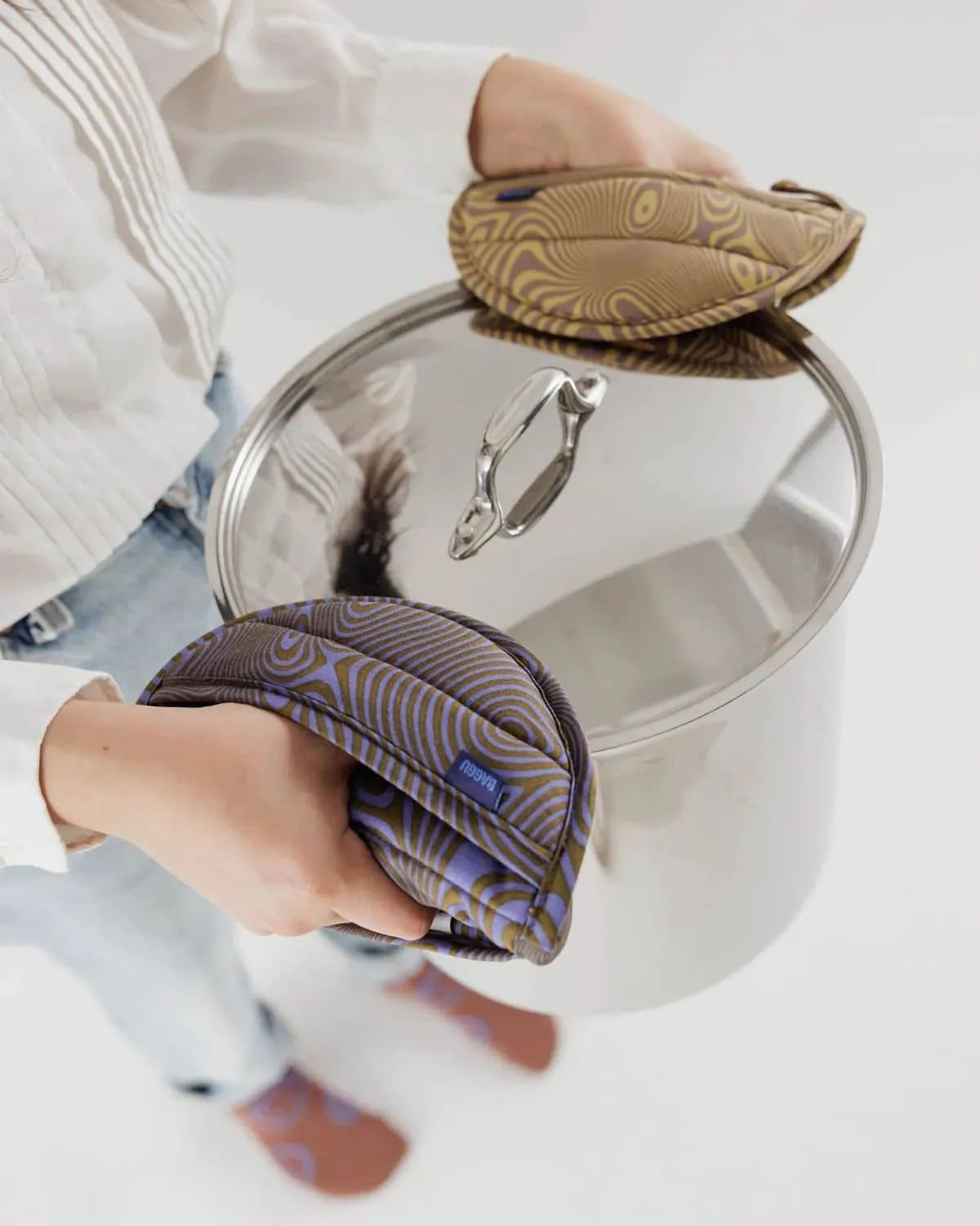 A person wearing a white shirt and blue jeans holds a large stainless steel pot by its lid and handle, using the BAGGU Pot Holder Set of 2 by BAGGU in brown and purple psychedelic patterned organic cotton.