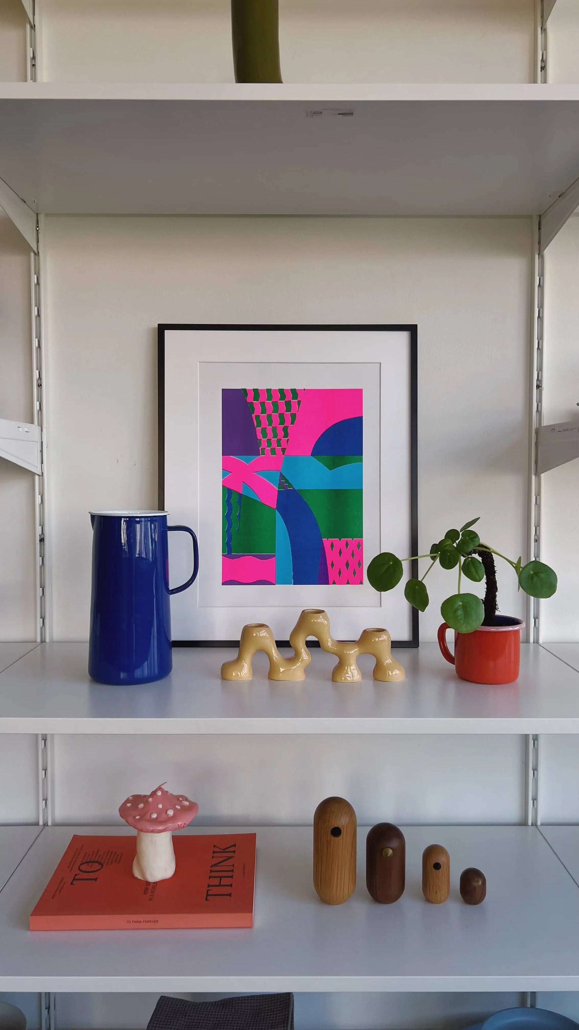 A minimalist shelf displays a Bbblob Freefall, 2022 Risograph print (30x42) with charming misregistration, a blue pitcher, a small potted plant in a red pot, a gold decor piece, a ceramic mushroom, and two small wooden vases.