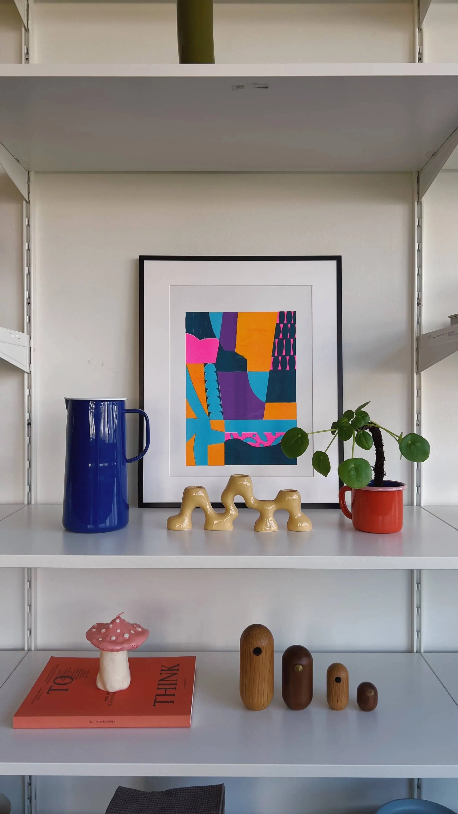 A white shelf displays a blue pitcher, Bbblobs Riding The Waves, 2022 Risograph (30x42), a small green plant in a red pot, a pink mushroom figurine, bird-shaped wooden objects, and a beige abstract sculpture.