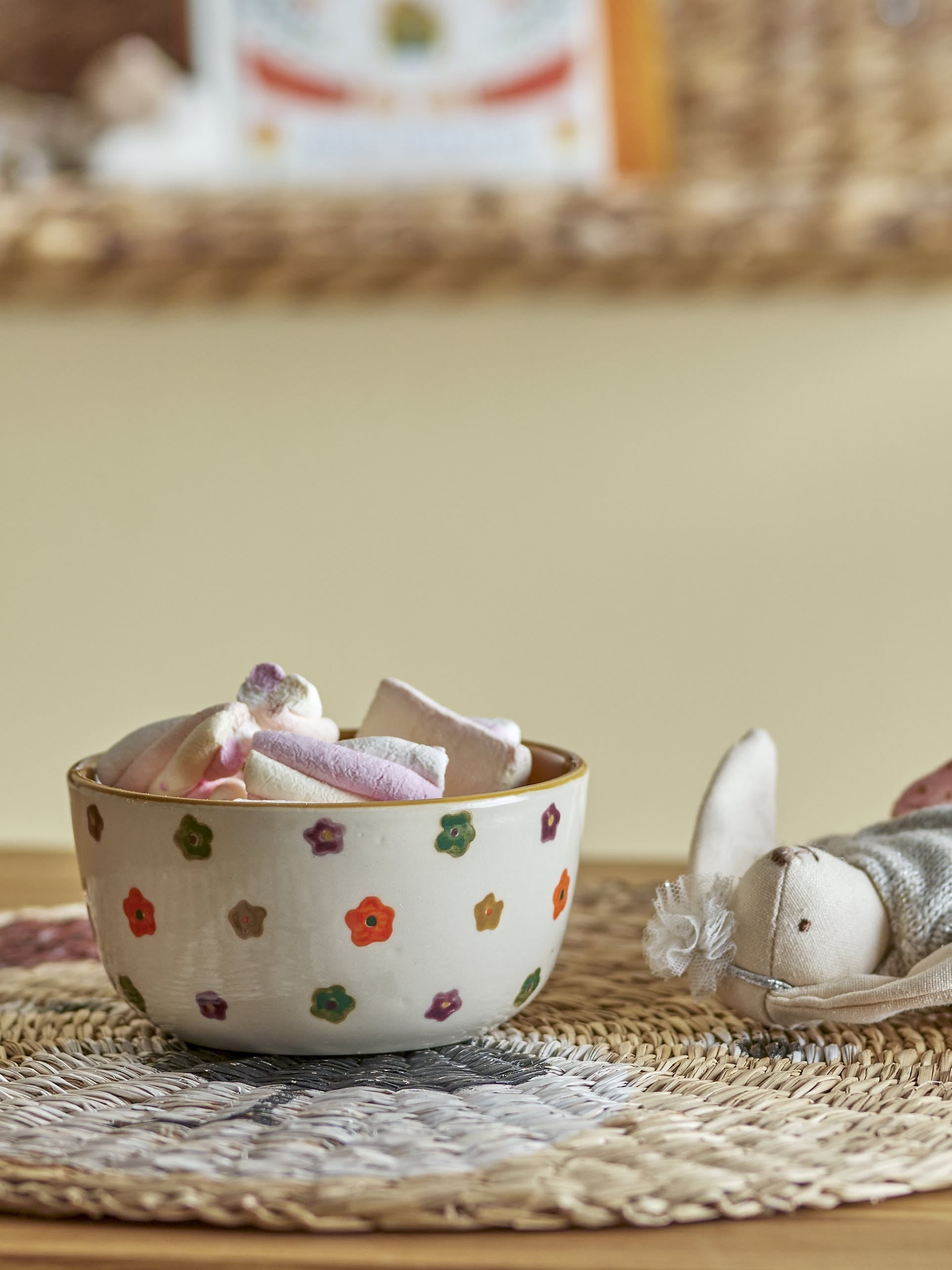 The Bloomingville Addy Bowl, featuring a hand-painted floral design, holds pastel marshmallows on a woven mat. Beside it is a soft bunny toy. The softly blurred background highlights the bowl’s charm. Brand: Bloomingville.