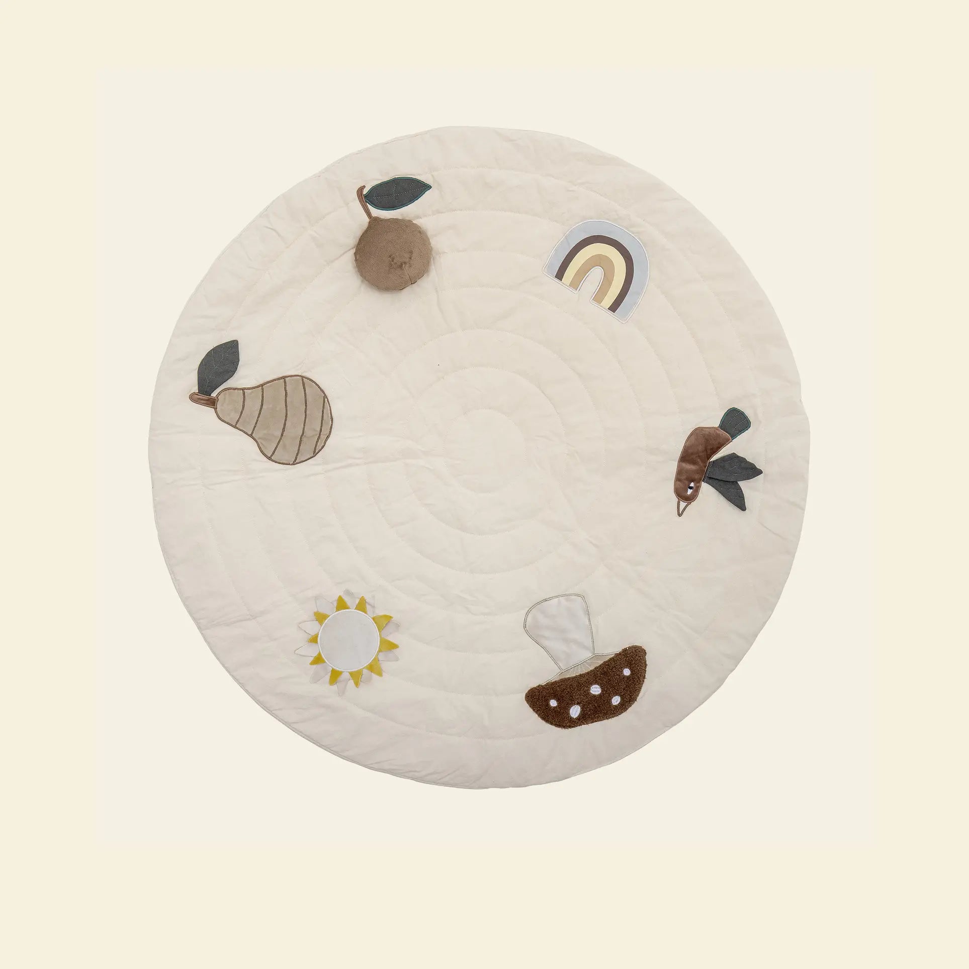The Bloomingville Agnes Play Mat features a round, cream-colored design with quilted circles and soft appliqués of a pear, apple, rainbow, fish, boat, and sun—ideal for sensory play and tactile exploration.