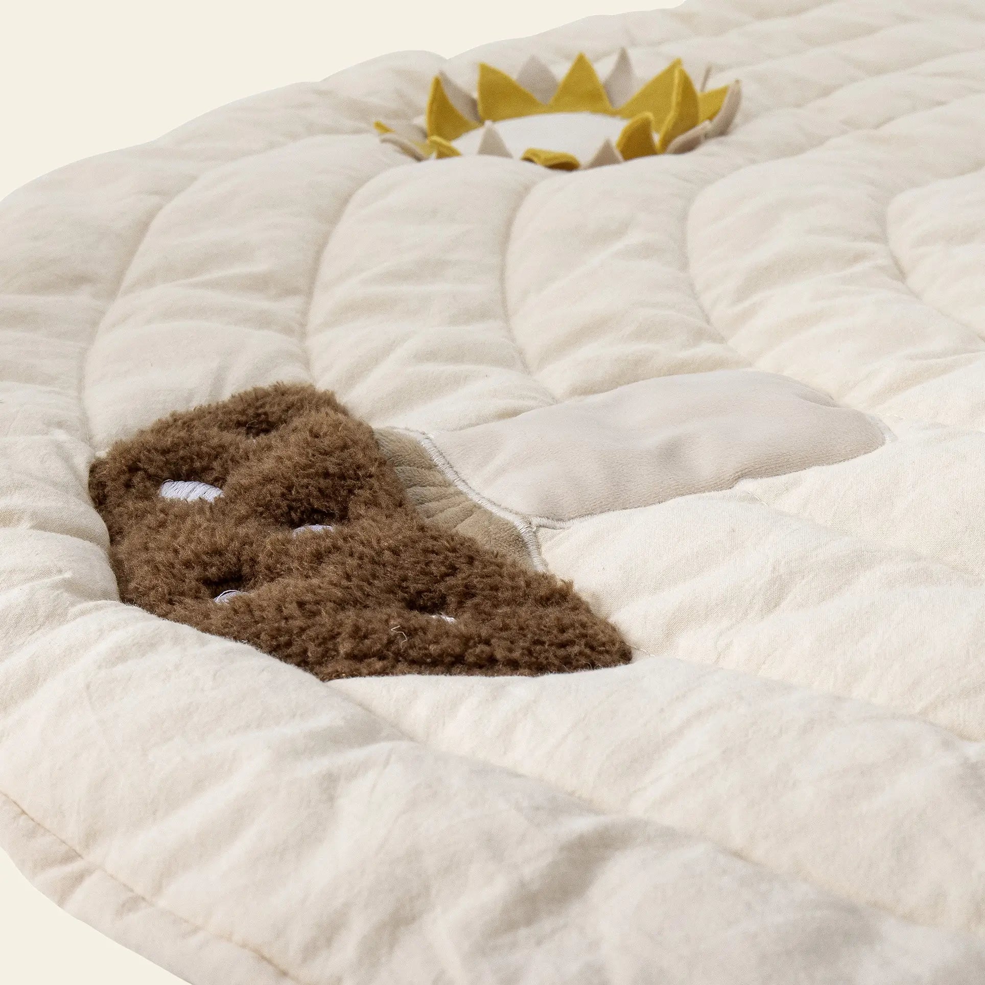 A close-up of the Bloomingville Agnes Play Mat by Bloomingville, featuring a soft cream quilted surface with fabric accents: a brown patch, light beige oval, and a sun-shaped yellow and white sensory element for tactile play.