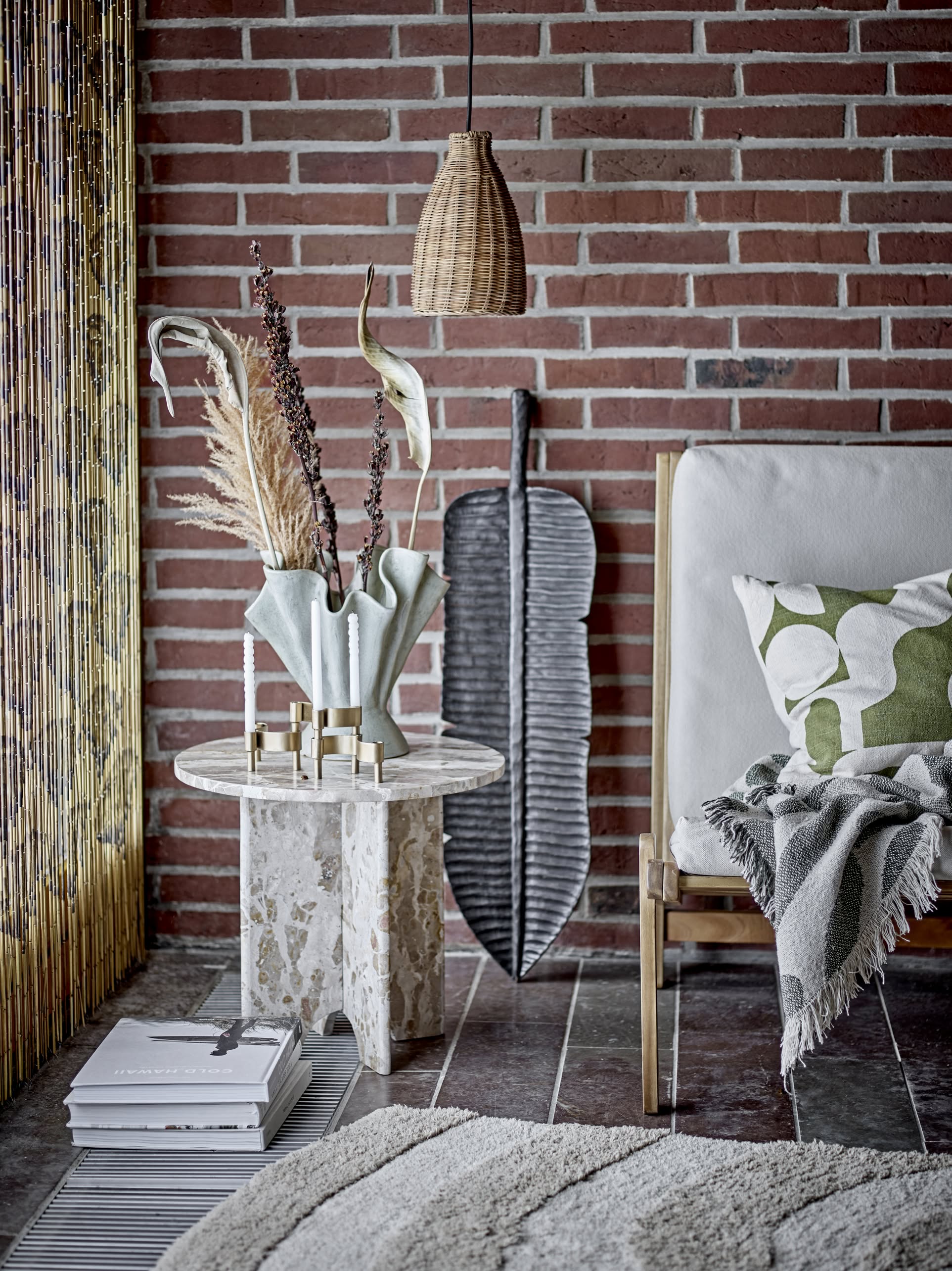 A cozy living space with a brick wall, a white cushioned chair, a green and white pillow, and the Bloomingville Alk Throw in recycled cotton. A marble side table with decor sits under a pendant lamp.