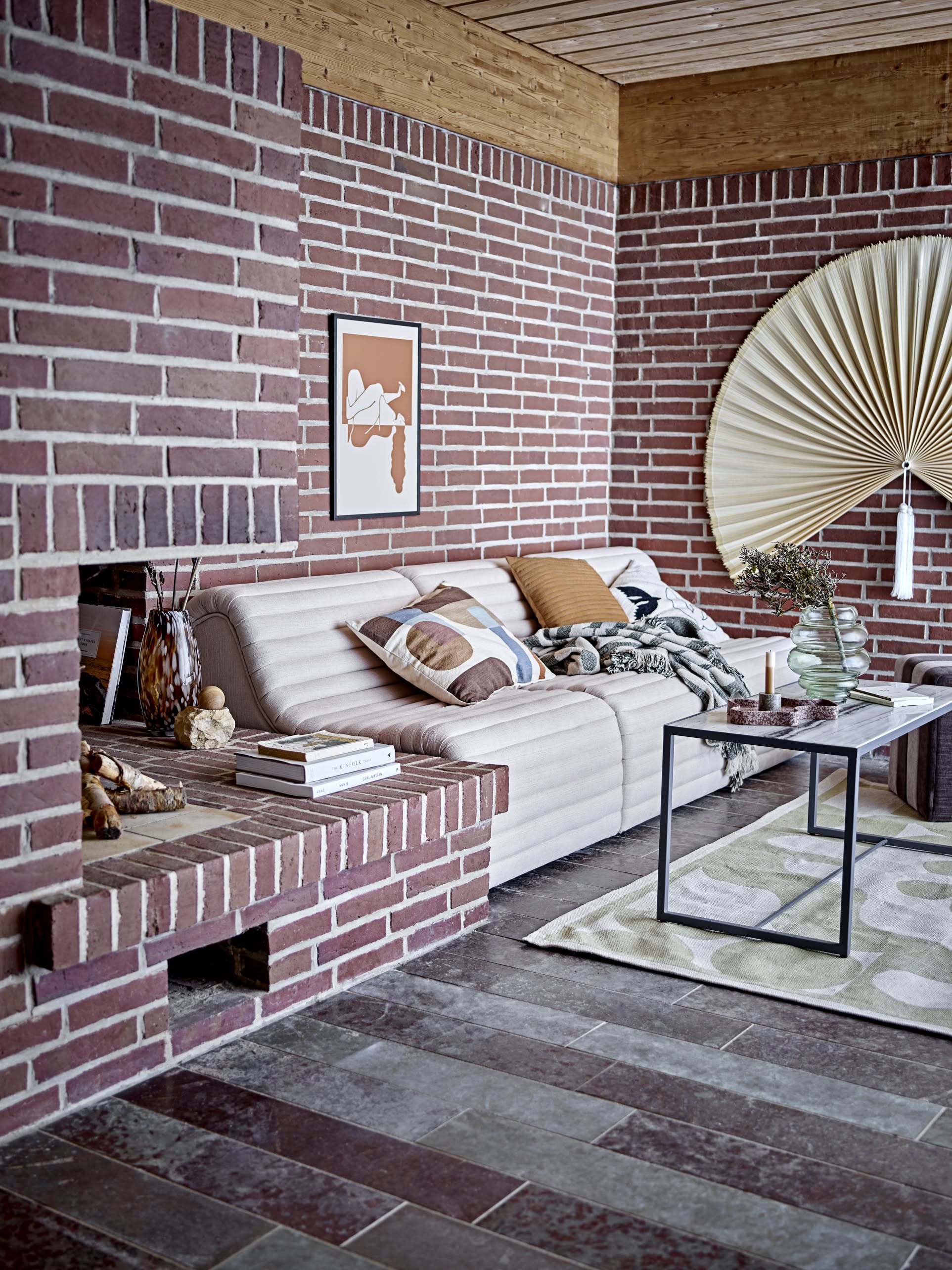 A cozy living room with red brick walls, a beige sofa topped with the Bloomingville Alk Throw, minimalist cushions, a modern coffee table, vases, artwork, and fan-shaped wall decor. Tiled floors and warm hues make the space inviting.