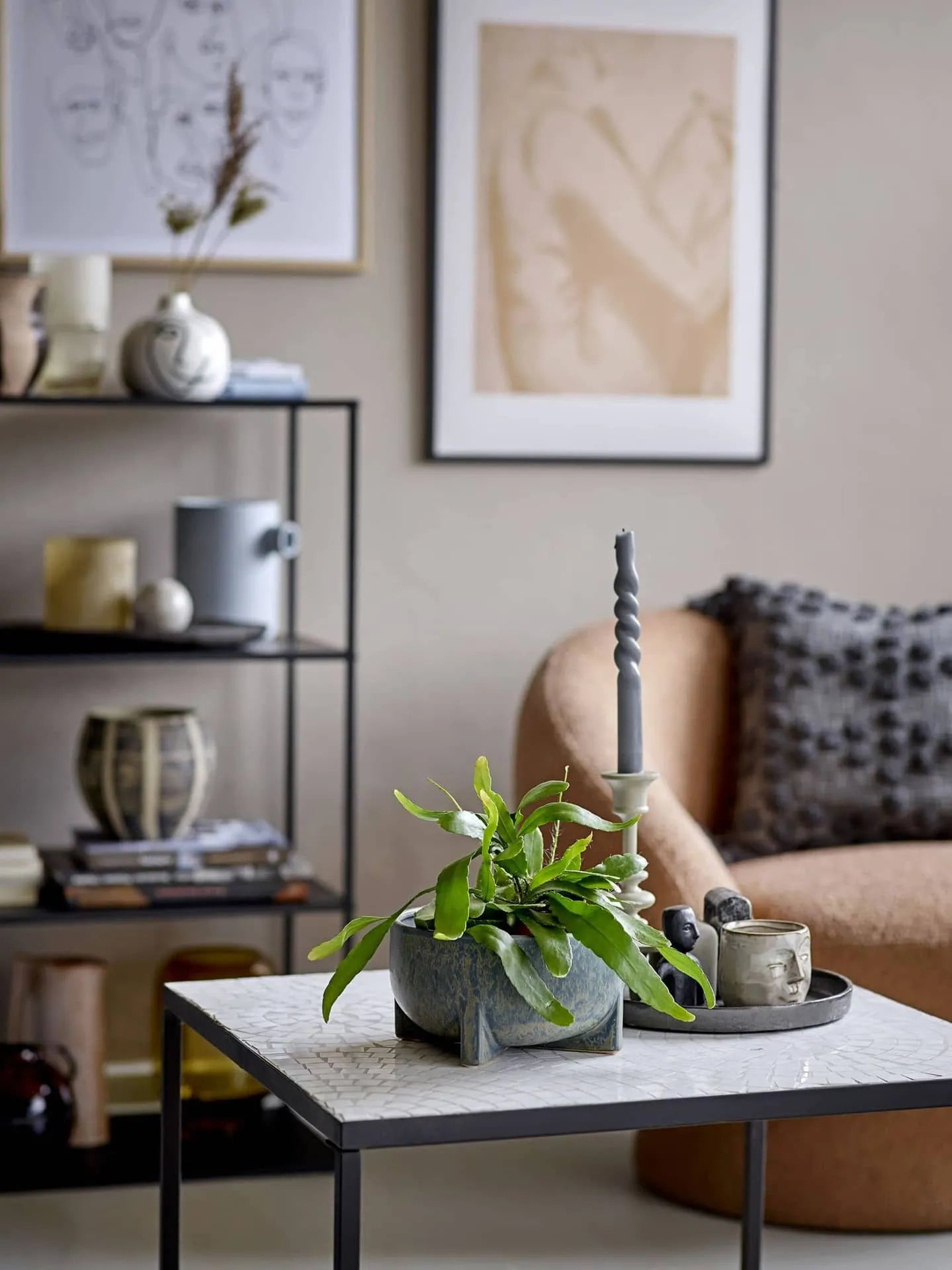 A modern living room features a beige armchair and a white tabletop with a Bloomingville Beryl Flowerpot filled with lush greenery, alongside a candle and mugs; shelves hold books, vases, decor, and abstract art decorates the wall.