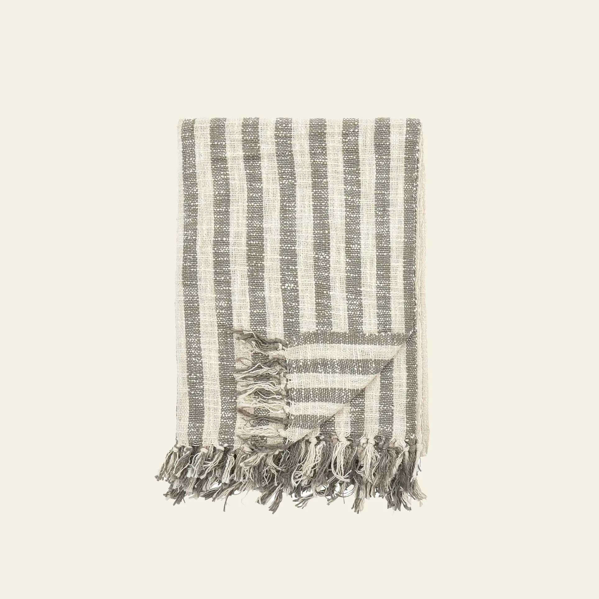 The Bloomingville Binnur Throw by Bloomingville is a folded, rectangular woven cotton blanket with vertical gray and off-white stripes, fringed edges, and a subtle texture, shown on a plain light background.