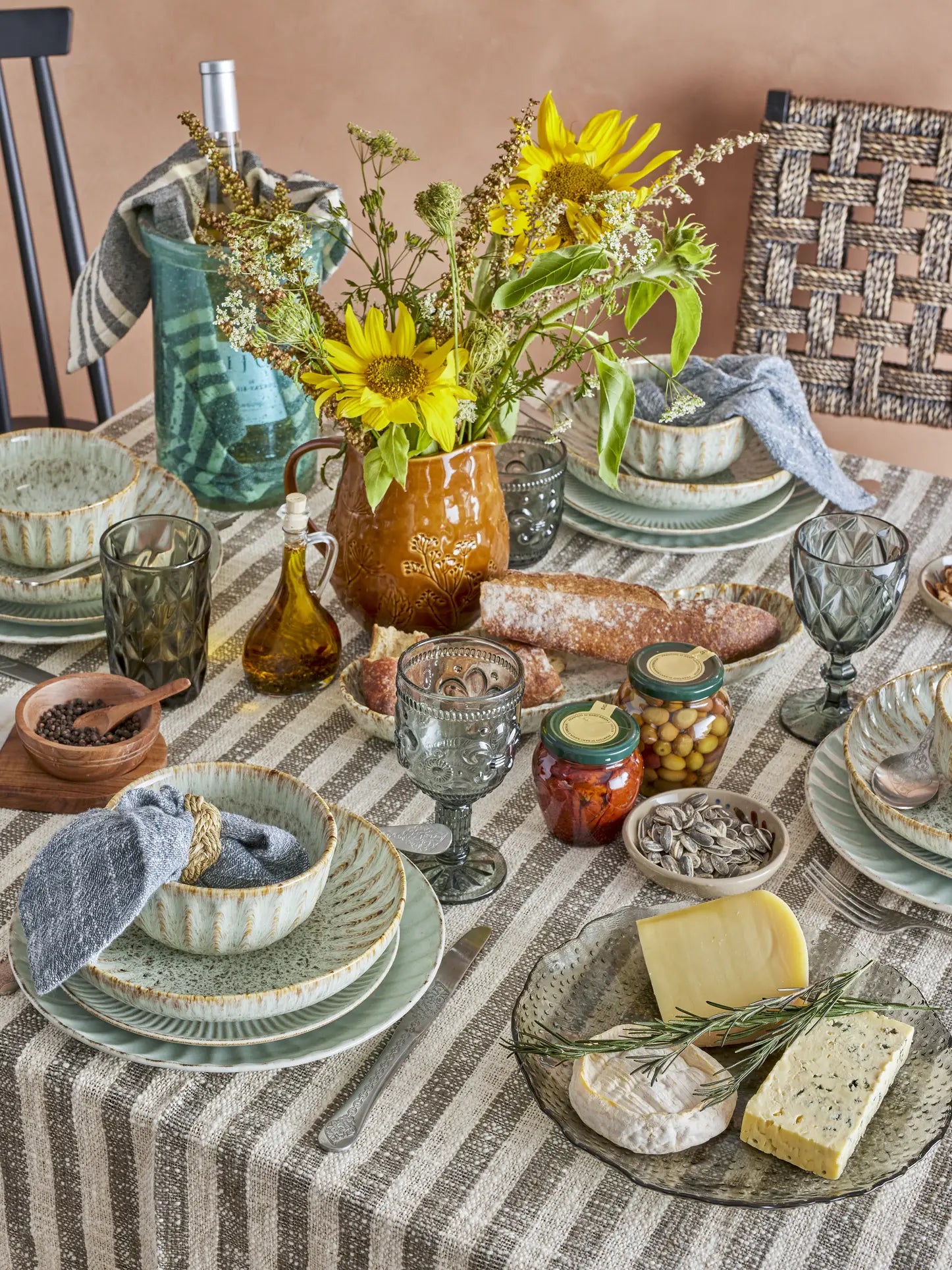 A cozy meal scene features a rustic table set with striped woven cotton linens, ceramic plates and bowls, vintage glasses, cheese, bread, olives, preserves, sunflowers in a pitcher—and the Bloomingville Binnur Throw by Bloomingville adds inviting texture.