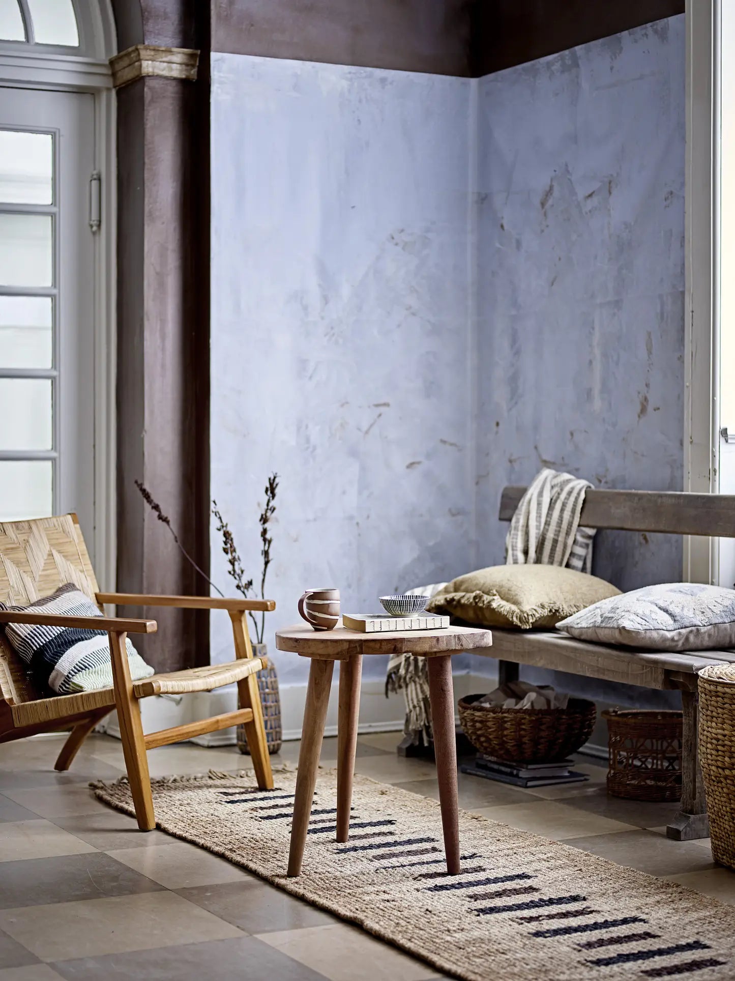 A cozy minimalist living space features a wooden armchair, small round table with a mug and book, woven cotton rug, and a bench topped with cushions and the Bloomingville Binnur Throw by Bloomingville against a light blue textured wall. Natural light fills the room.
