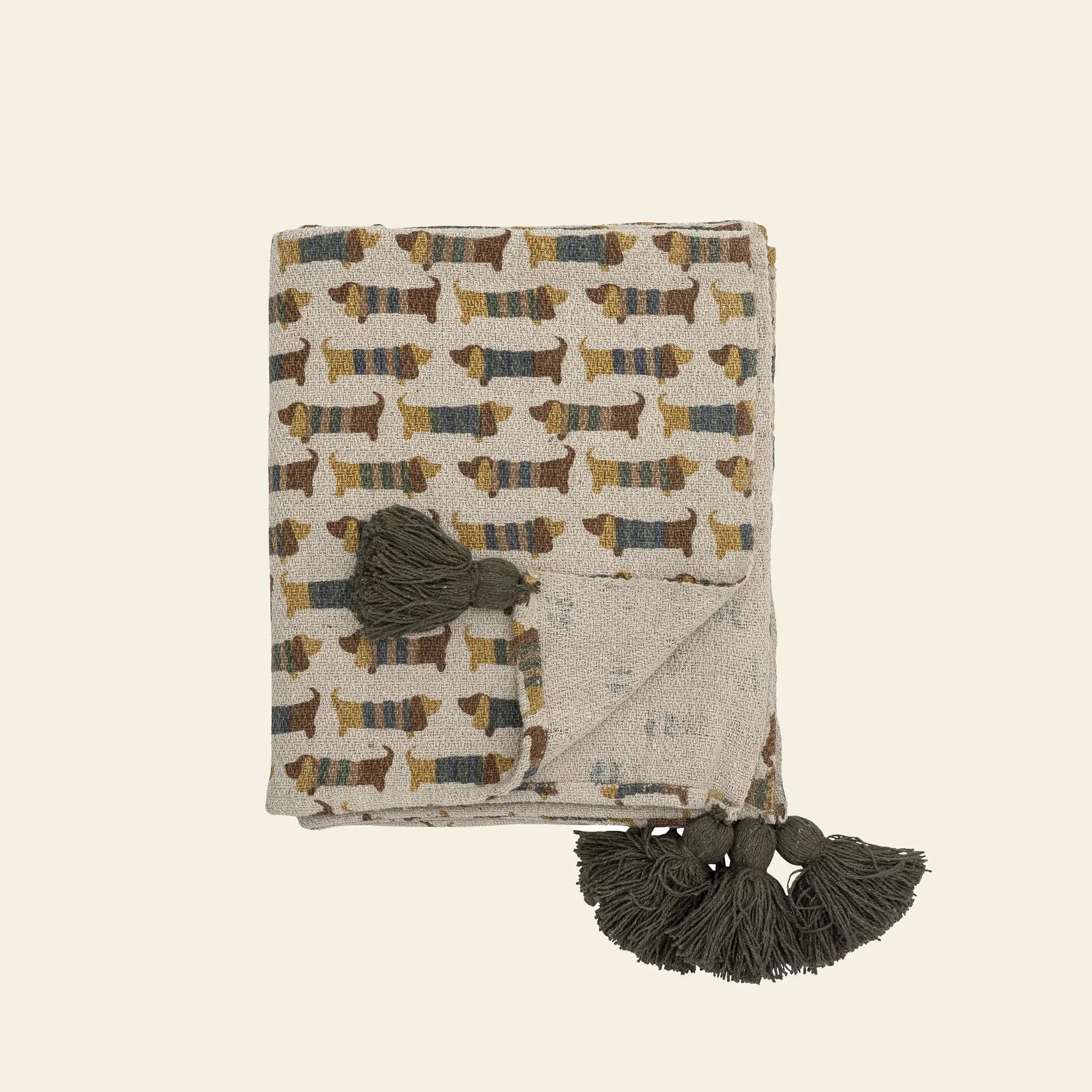 The Bloomingville Charlie Throw by Bloomingville is a folded recycled cotton blanket featuring a dachshund pattern in earthy tones, three dark green tassels on one corner, and a plain light background.