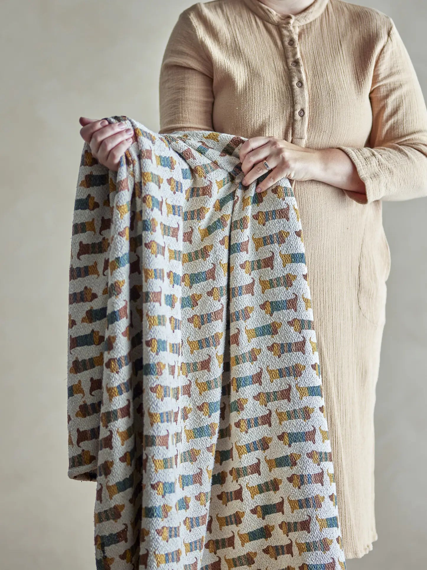 A person in a beige, long-sleeved dress holds the Bloomingville Charlie Throw by Bloomingville, a recycled cotton blanket with rows of abstract, multicolored dog shapes on a light background. The persons face is not shown.