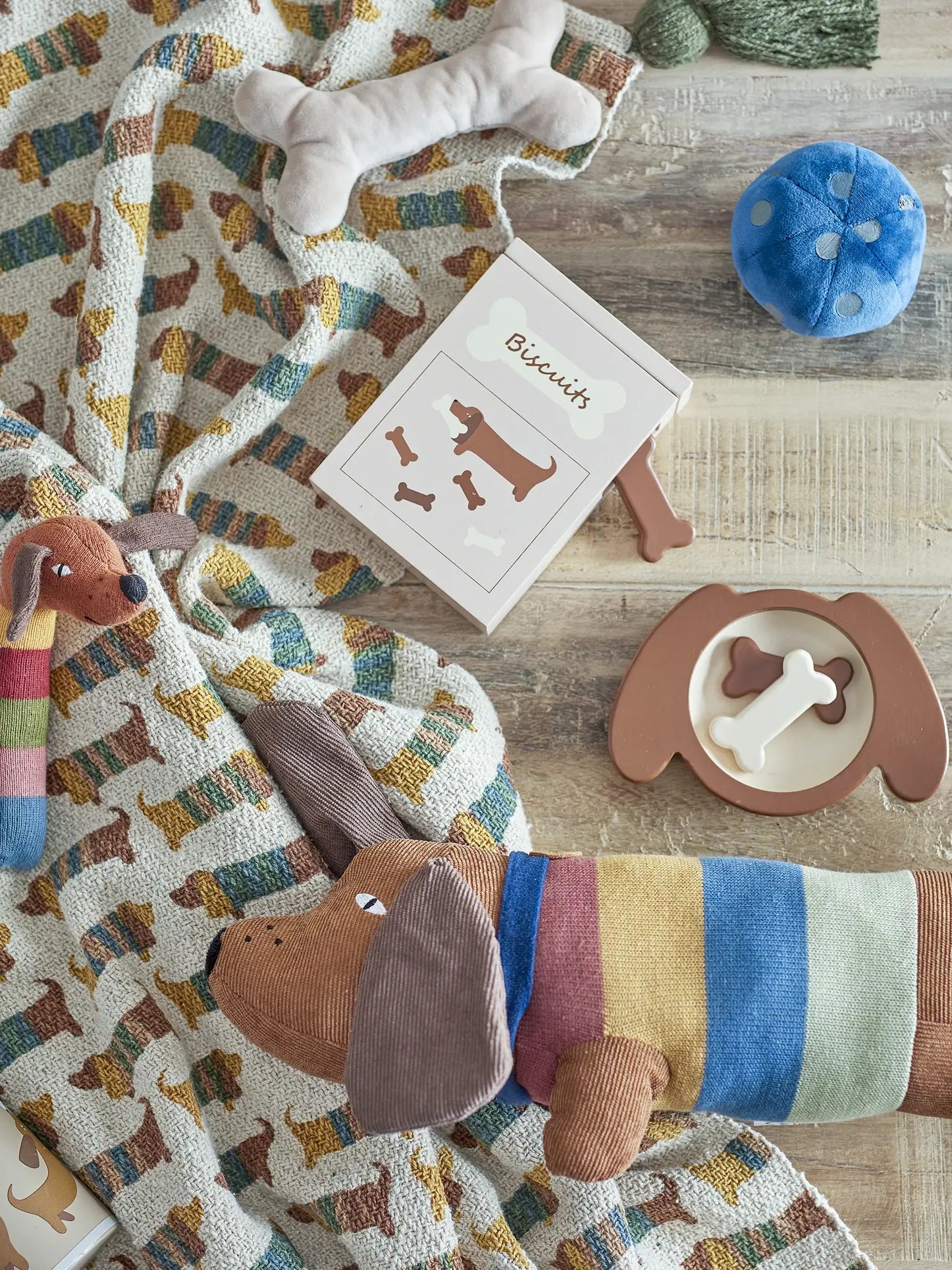 A cozy scene with dog-themed toys, a plush pup in a rainbow sweater, a Biscuits book, and the Bloomingville Charlie Throw by Bloomingville made from recycled cotton spread across the wooden floor.