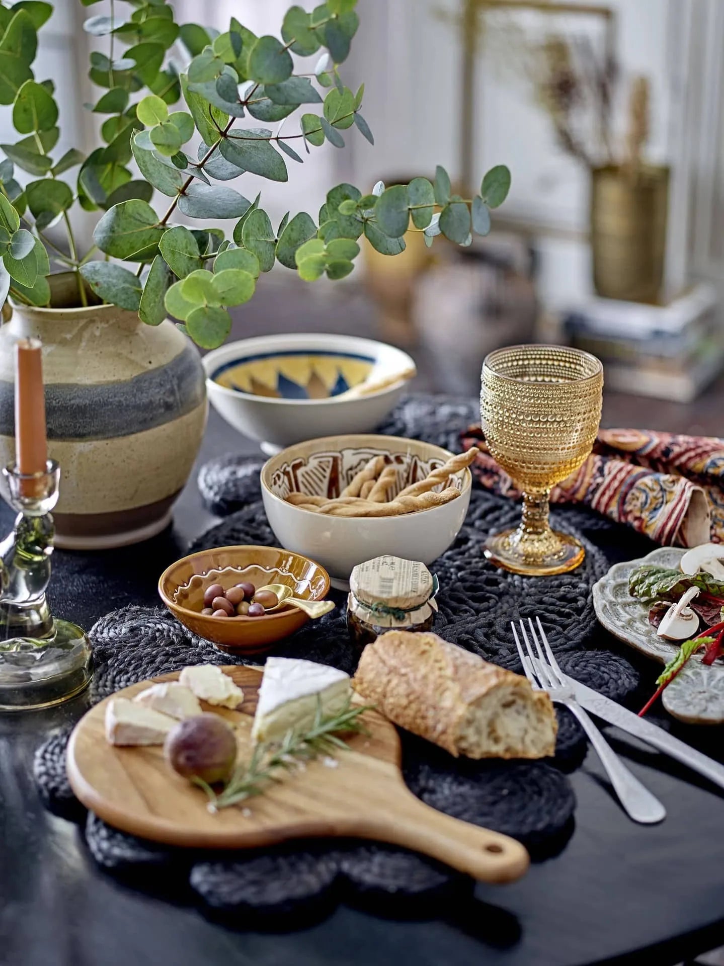A cozy, inviting kitchen scene is set with a stylish table featuring a vase of eucalyptus, candle, patterned snack dishes, glass goblet, and cheese and bread on the Bloomingville Dana Cutting Board by Bloomingville.