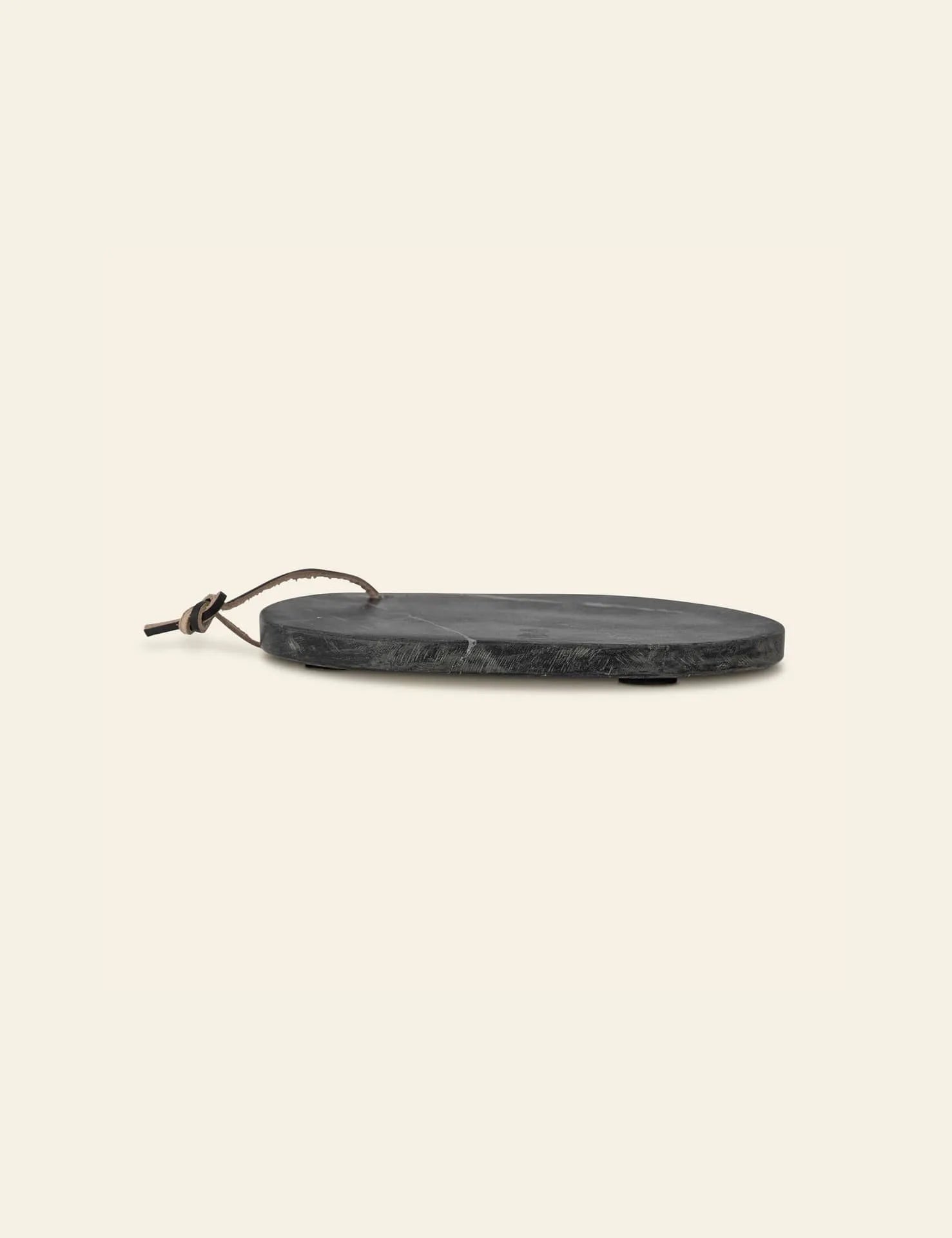 The Bloomingville Daris Cutting Board is a round, flat black slate board with a brown leather loop, shown from the side on a beige background. It features a smooth cutting surface ideal for stylish serving.