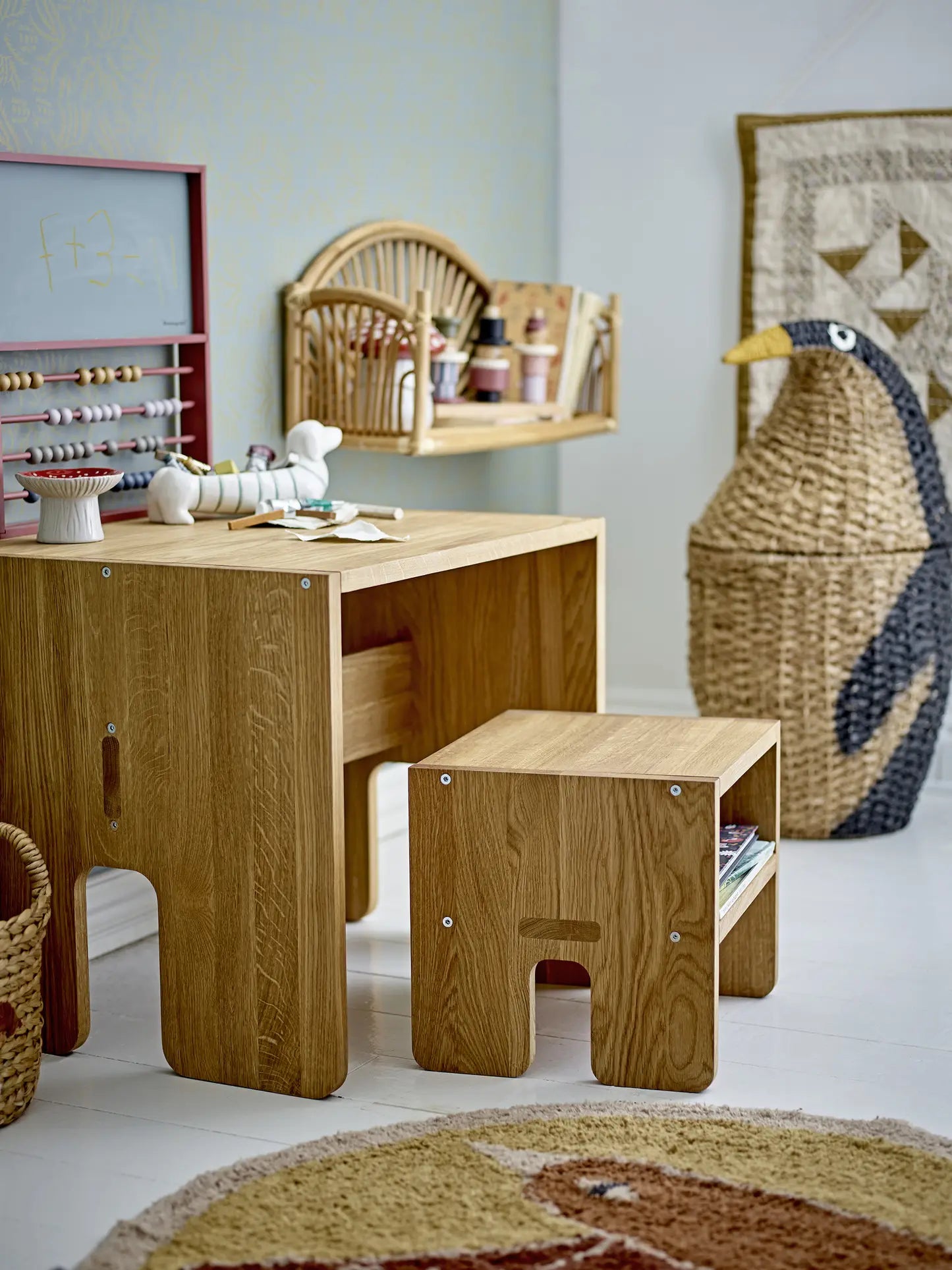 A children’s playroom features a Bloomingville Djaco Bowl, a wooden table with matching stool, a large woven basket for storage, wall rack with art supplies, an abacus, toys, and an animal-themed round rug.