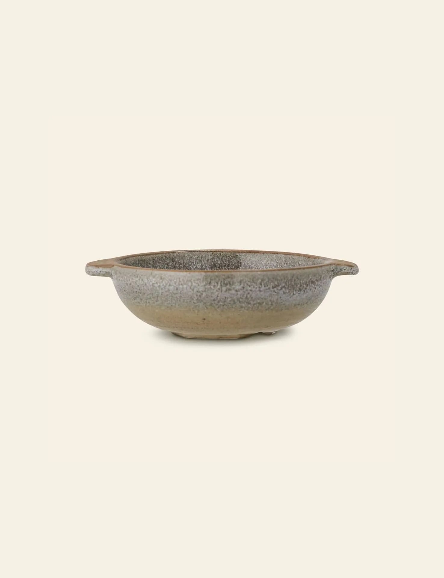 The Bloomingville Hariet Bowl by Bloomingville is a shallow, round stoneware bowl with small side handles and a speckled gray and beige glaze. Handcrafted details bring subtle charm to this piece, shown against an off-white background.