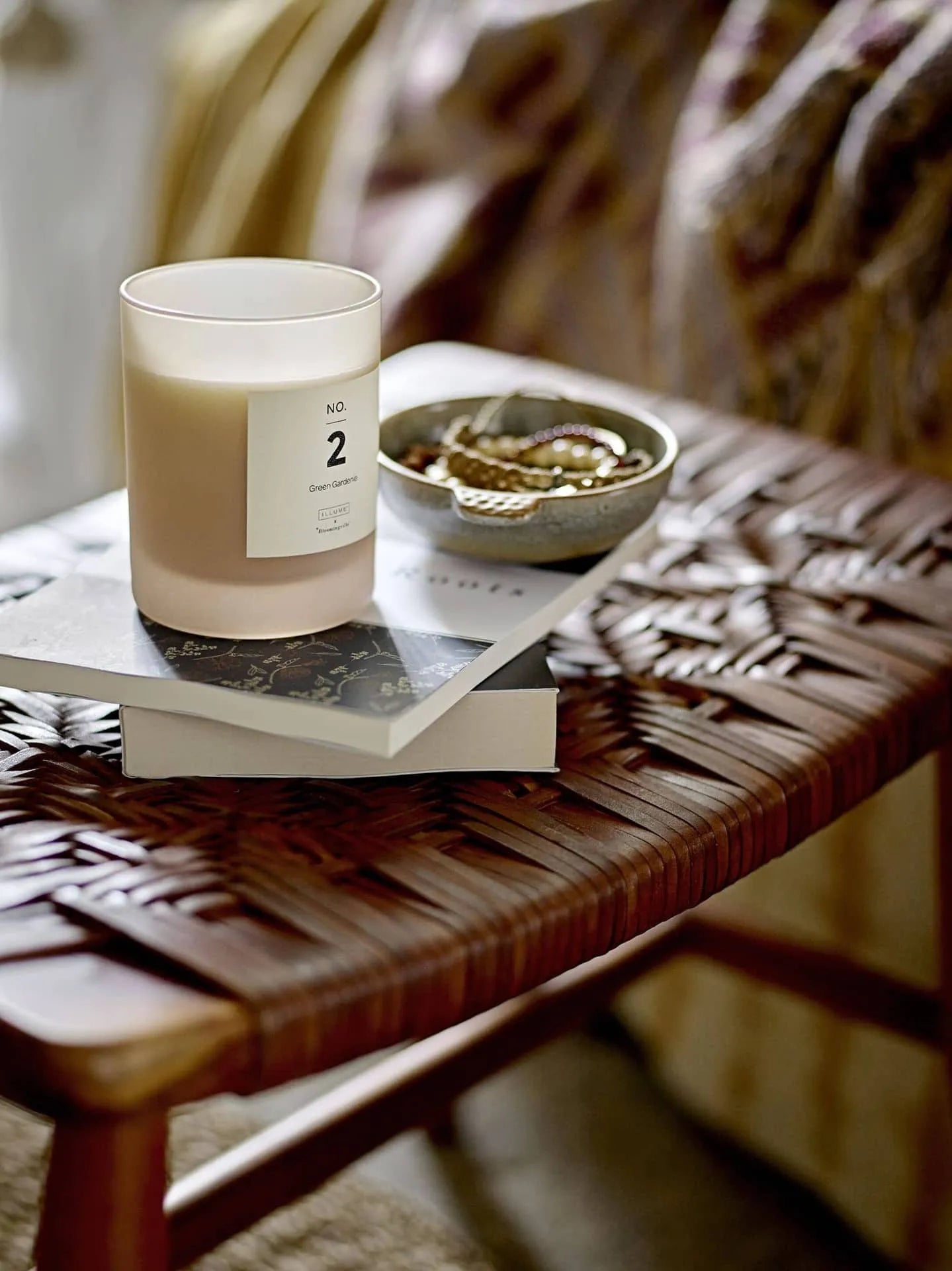 A white candle labeled “No. 2” sits on stacked books atop a woven rattan side table. Next to it, jewelry rests in the Bloomingville Hariet Bowl by Bloomingville, with patterned fabric visible in the background.
