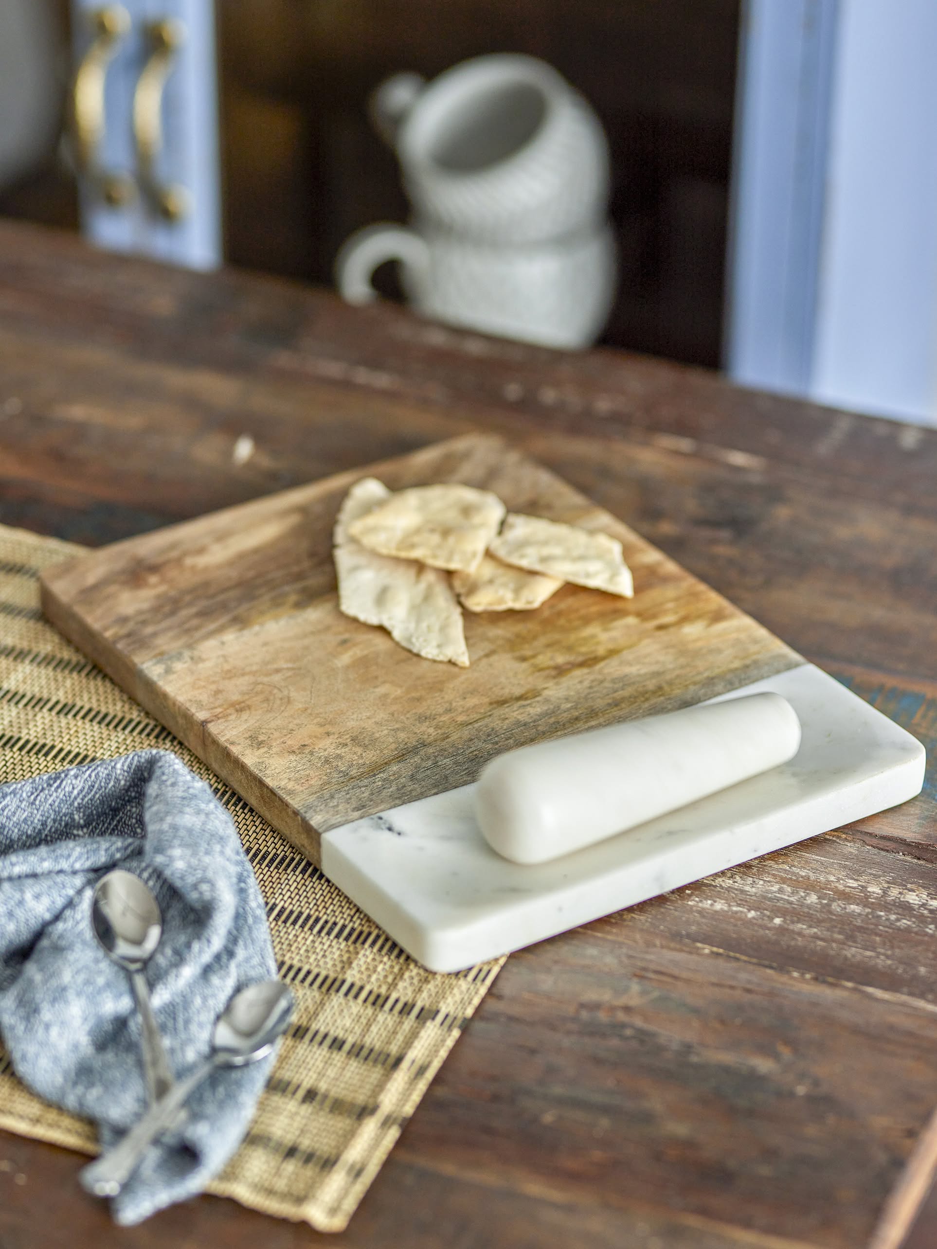 The Bloomingville Havannah Cutting Board with Pestle by Bloomingville is shown on a woven placemat, topped with broken flatbread and a white marble rolling pin. Two spoons and a gray napkin sit beside it on a rustic wooden table.