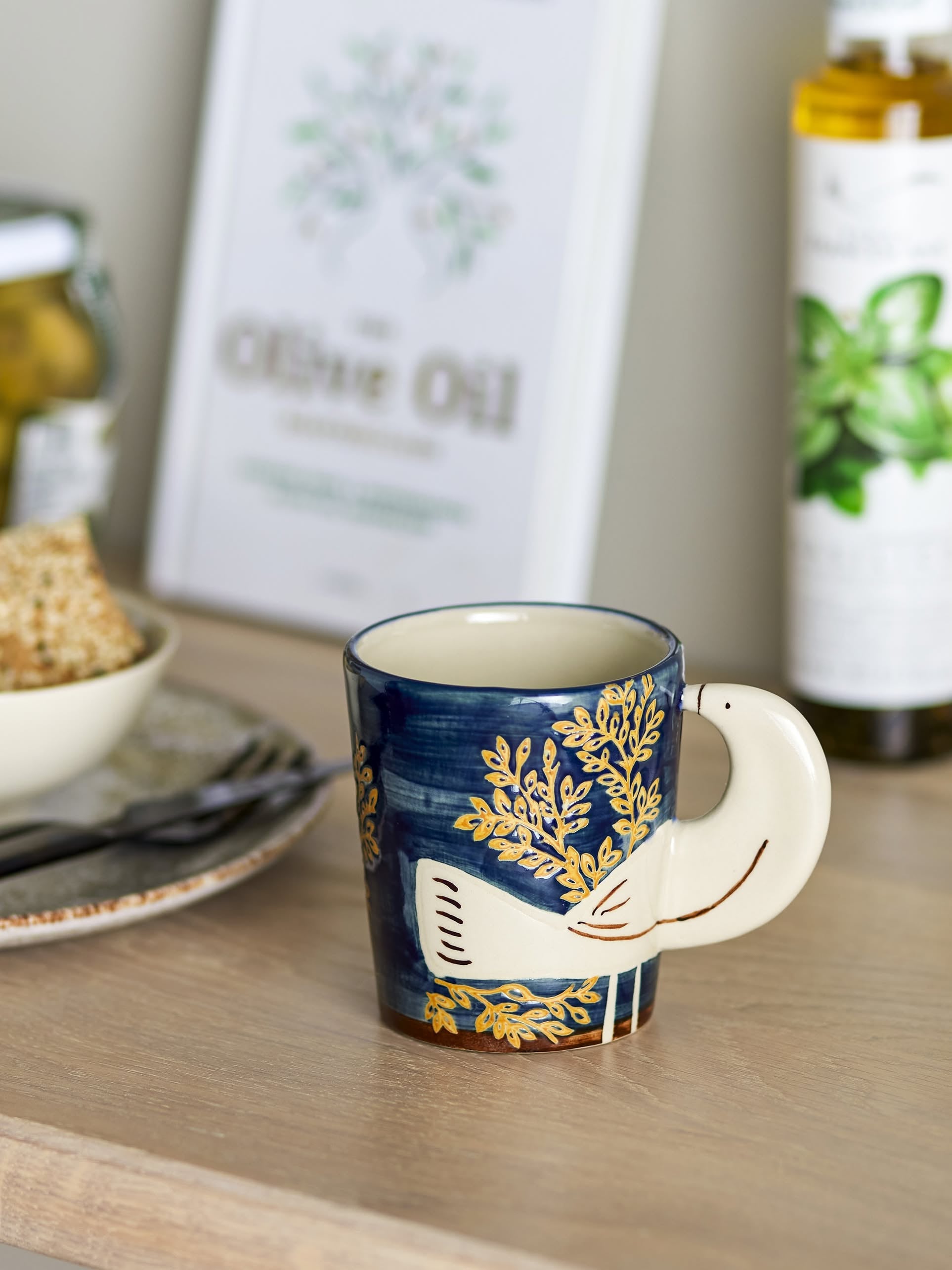 The Bloomingville Hezha Cup - Blue, featuring a hand-decorated bird motif and gold leaf design by Bloomingville, sits on a wooden table alongside bread, a jar, and olive oil.