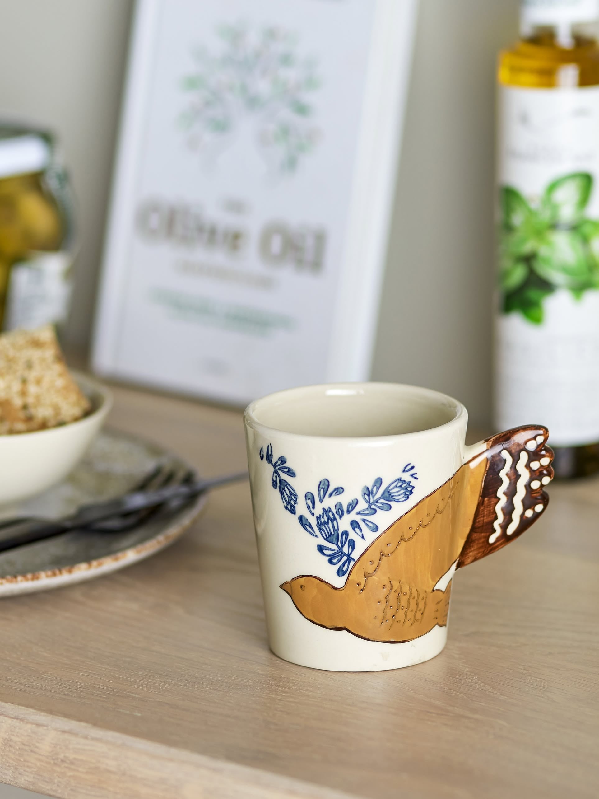 The Bloomingville Hezha Cup - Nature, a ceramic mug with a bird motif and sculpted wing handle by Bloomingville, sits on a wooden surface near bread, a fork, an olive oil bottle, and a framed print in the background.