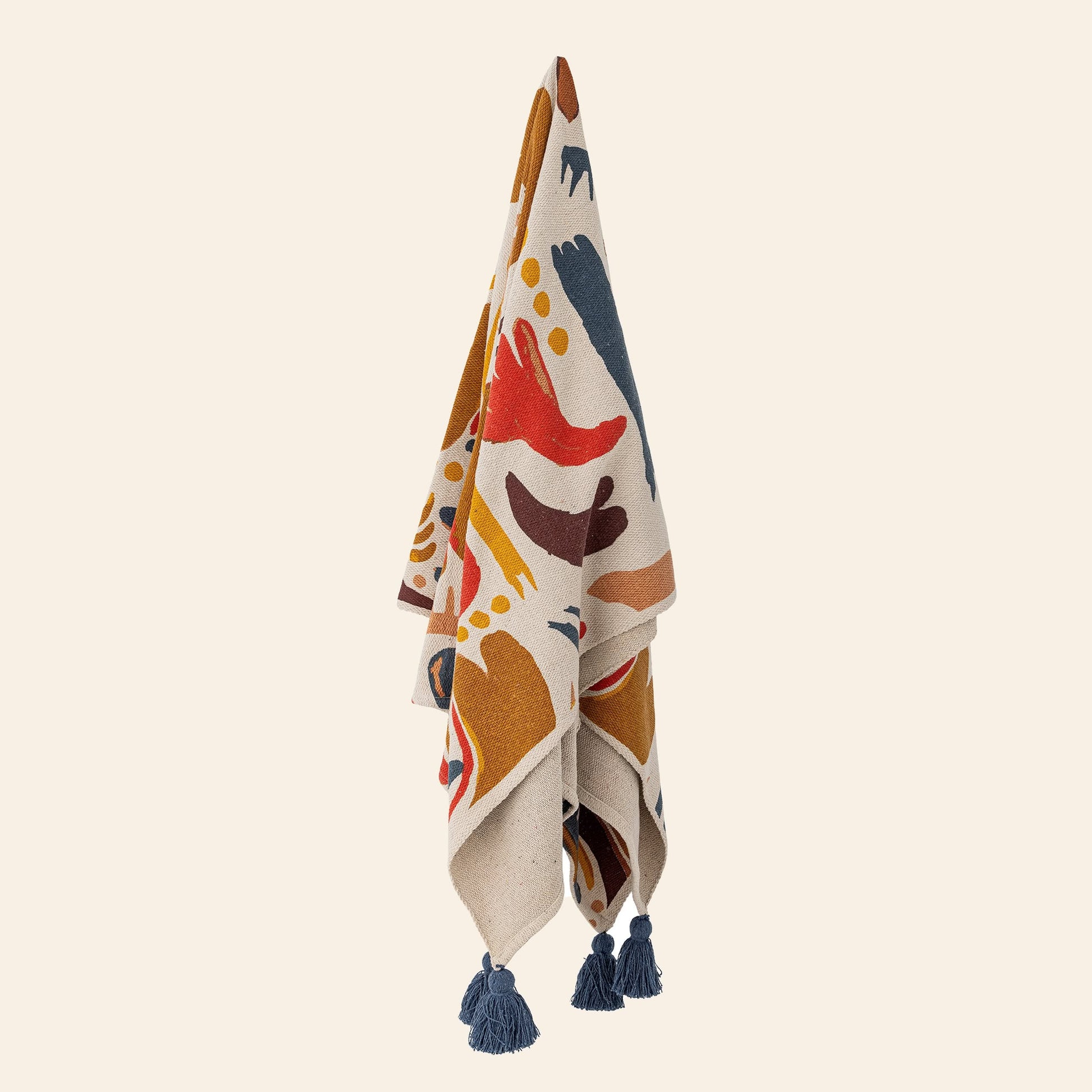 The Bloomingville Horus Throw, a recycled cotton blanket with abstract red, blue, mustard, and beige patterns plus blue tassels, hangs vertically against a plain beige background.