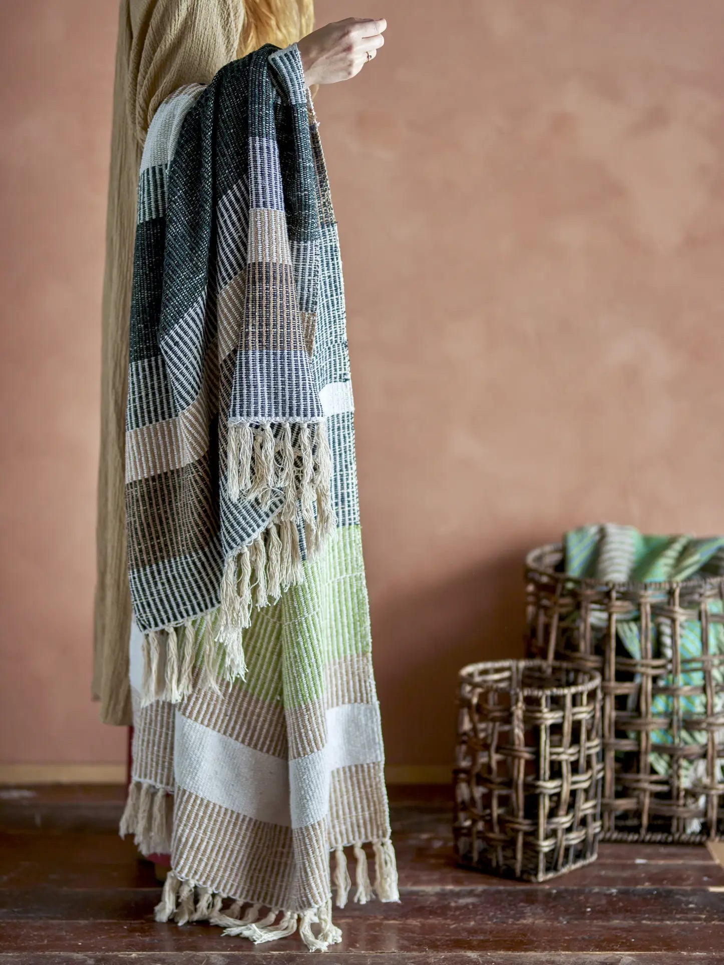 A person holds a Bloomingville Isnel Throw, a multicolored woven blanket with stripes and fringe. Another recycled cotton throw from Bloomingville drapes over a wicker basket on a wooden floor against a warm beige wall.