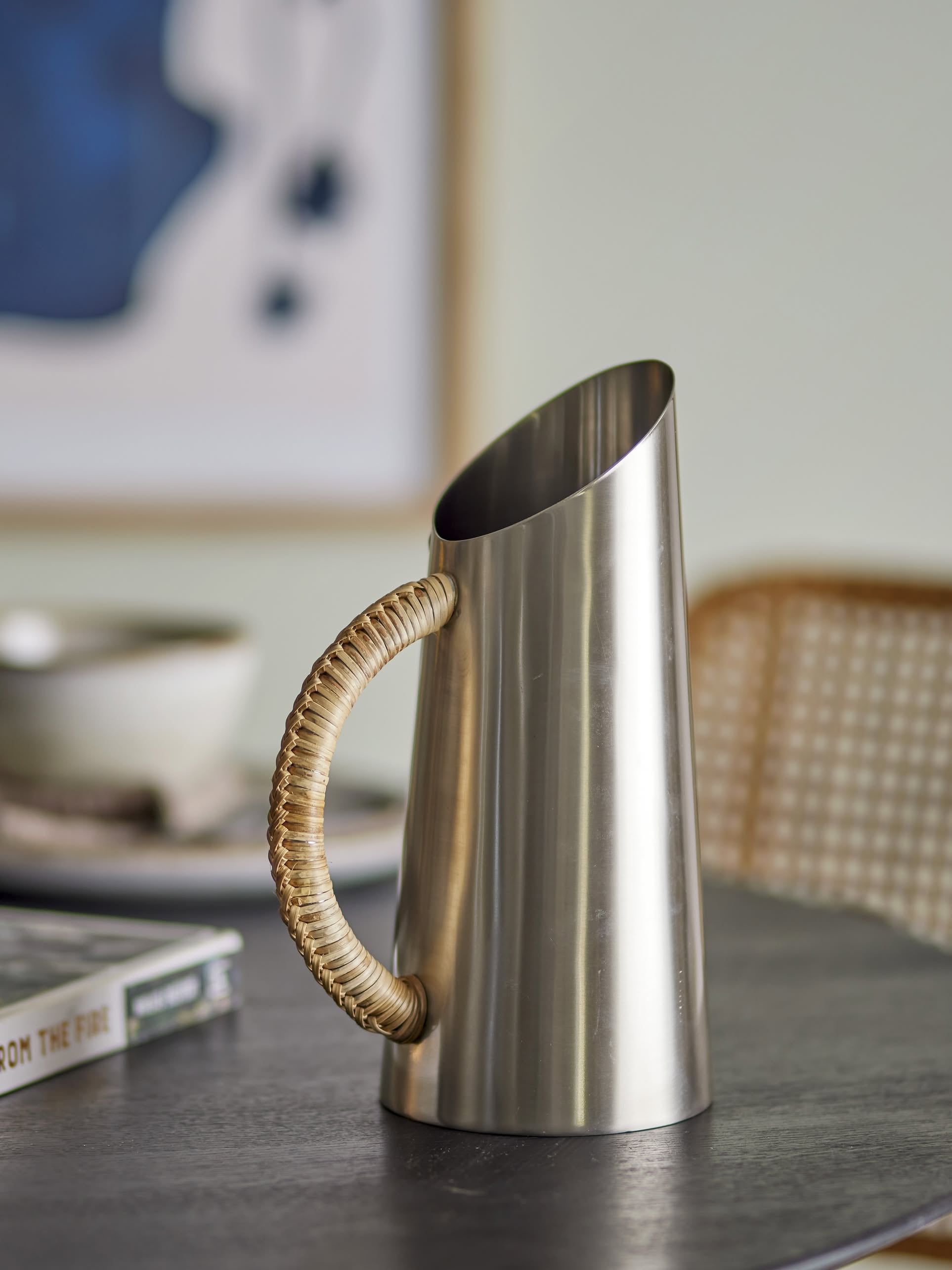 The Bloomingville Juanita Jug by Bloomingville, featuring stainless steel and a rattan-wrapped handle, rests on a dark tabletop. A blurred chair, book, and abstract artwork in the background add to the contemporary décor.