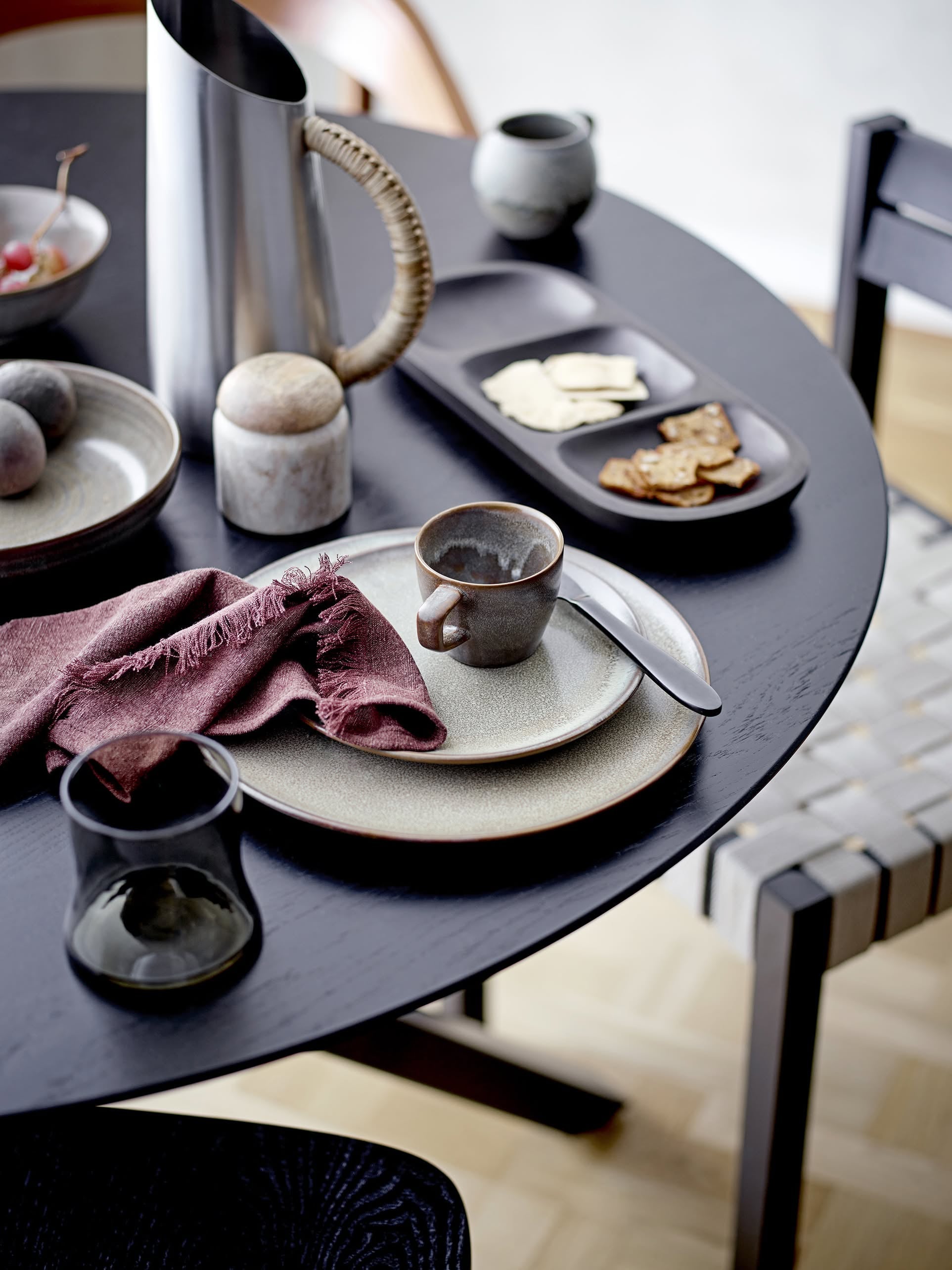 A dark round table set with a ceramic plate, cup, spoon, folded maroon napkin, glass, a bowl of fruit, salt or pepper shaker, trays with crackers and cheese in contemporary style, and the Bloomingville Juanita Jug by Bloomingville.
