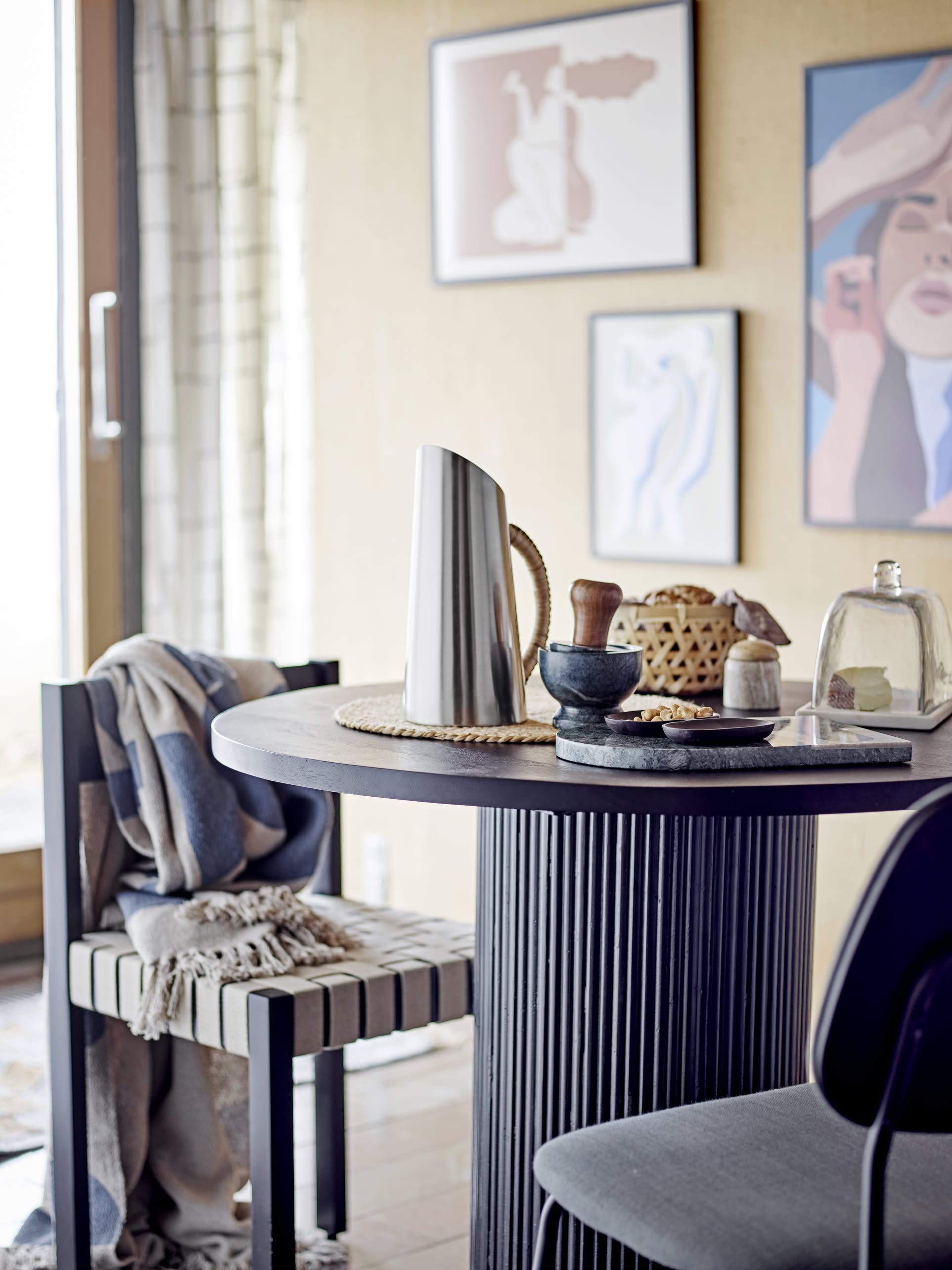A modern round dining table with the Bloomingville Juanita Jug, mortar and pestle, covered plate, and basket sits by a window. A striped chair with a draped blanket is nearby amid art prints on a beige wall.