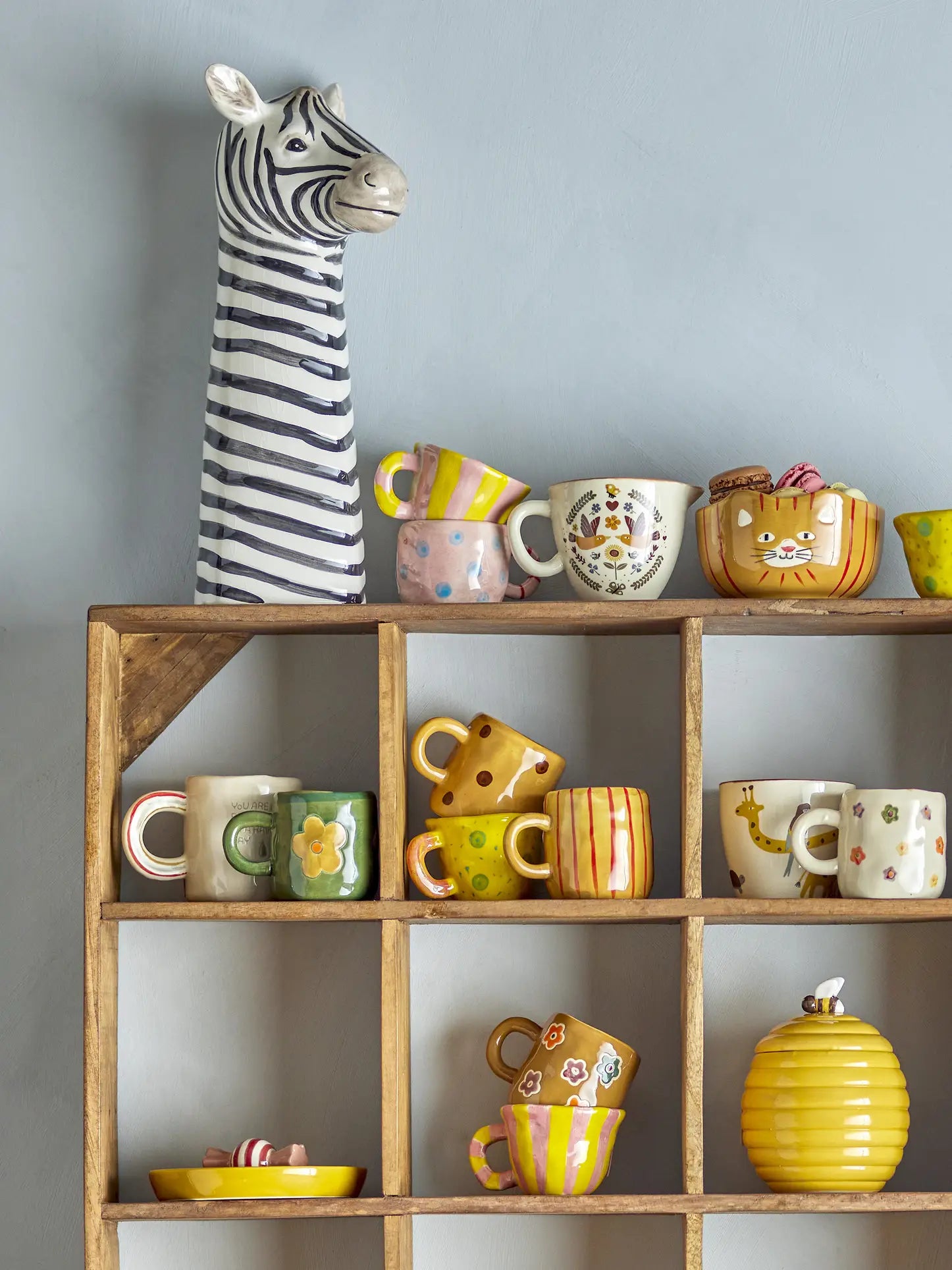 A decorative shelf displays colorful mugs and bowls. On top is a ceramic zebra, surrounded by mugs and a bowl from the Bloomingville Kittie Tableware by Bloomingville, featuring cat motifs and vibrant floral patterns.
