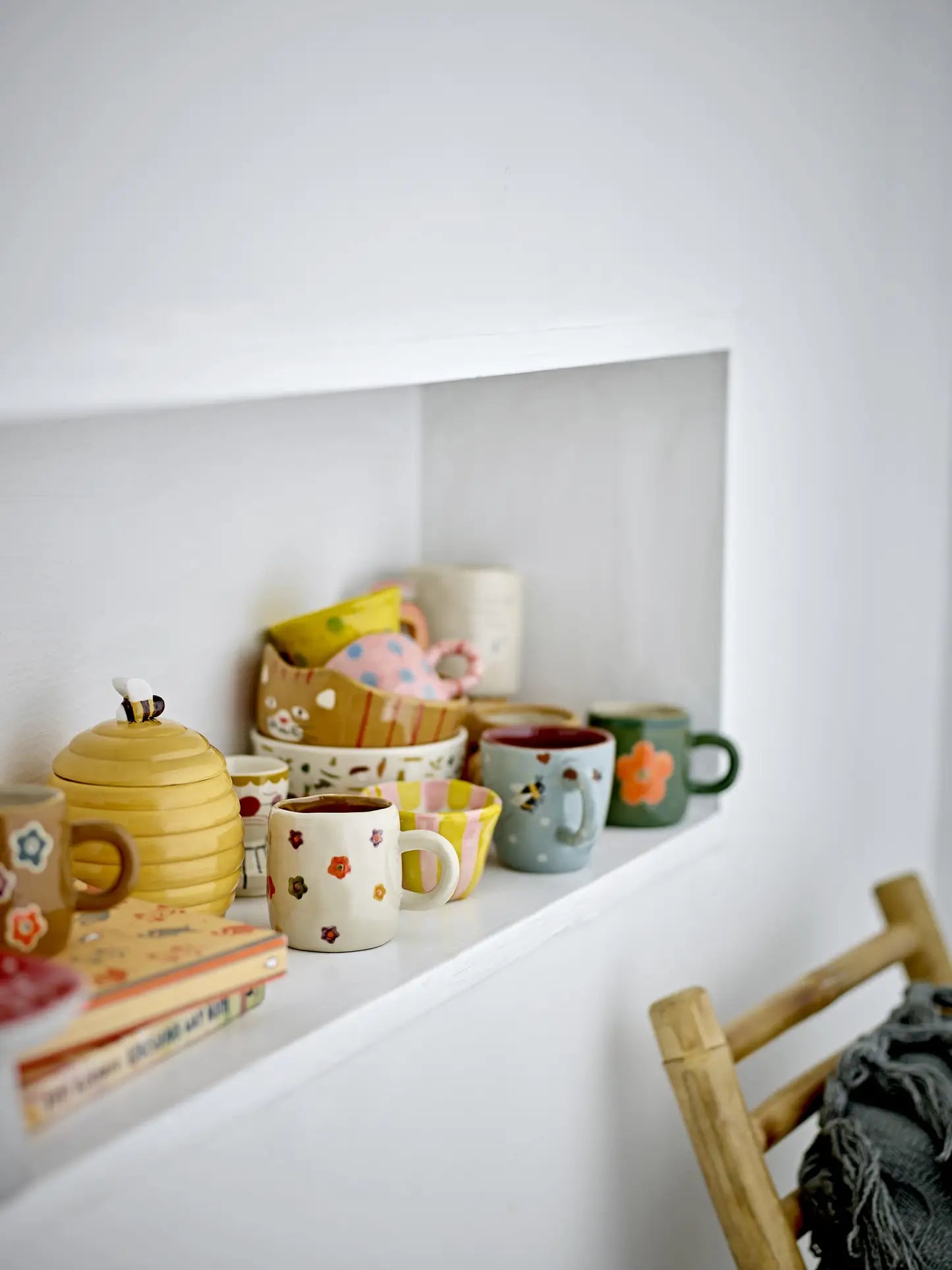 A white shelf inset in a wall holds colorful mugs, bowls, the Bloomingville Kittie Tableware featuring playful cat motifs, a beehive-shaped container, and stacked books. The edge of a wooden chair draped with a gray blanket appears in the foreground.