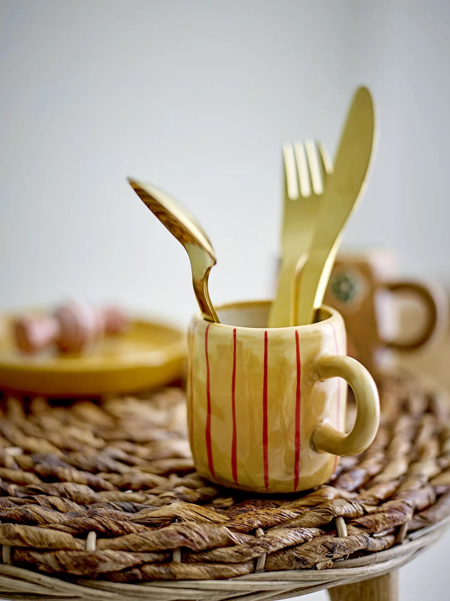 A yellow ceramic mug with red stripes from the Bloomingville Kittie Tableware holds a gold spoon, fork, and knife on a woven placemat. A matching plate and another mug from Bloomingville are blurred in the background.