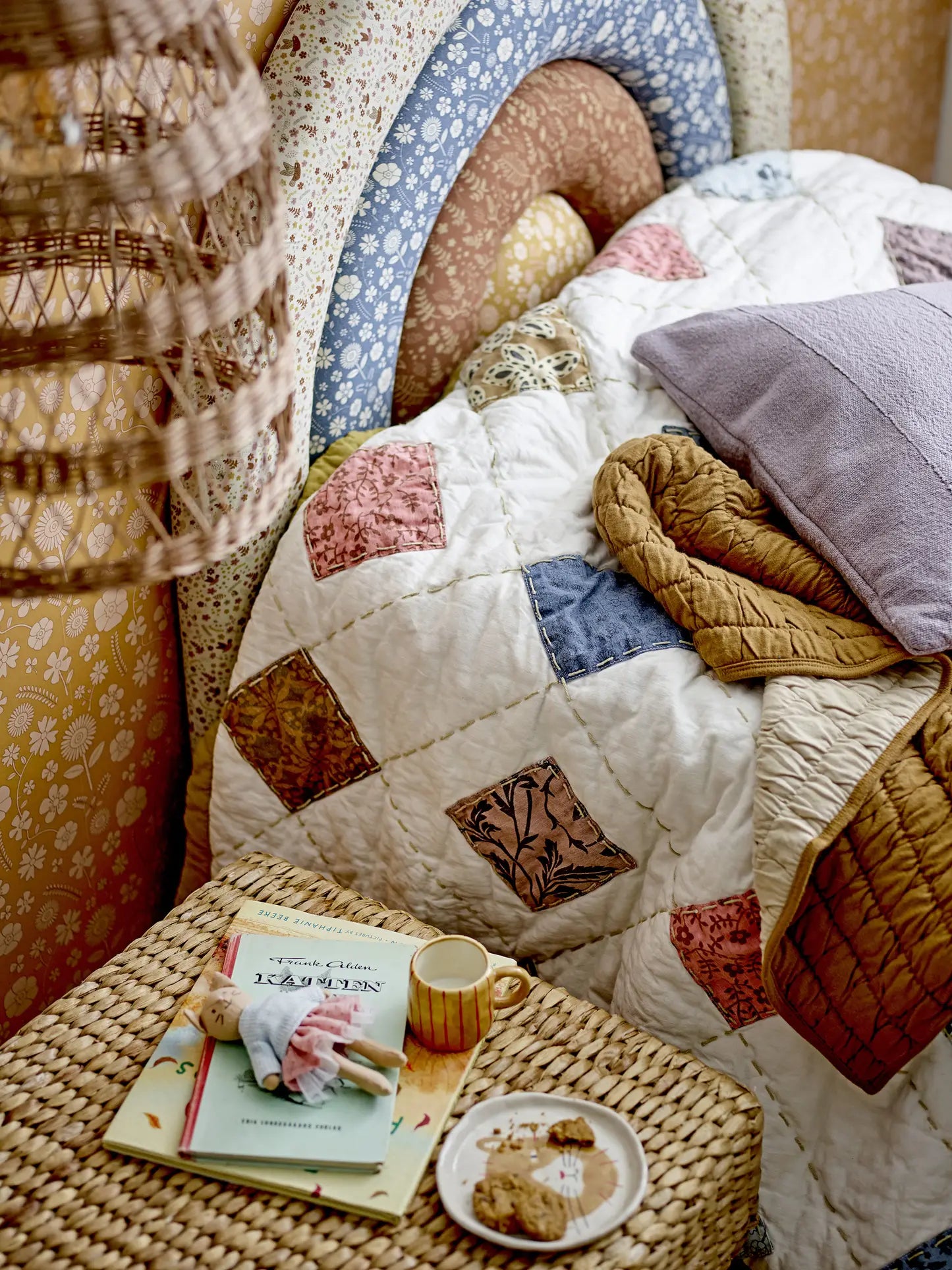 A cozy bed with a patchwork quilt and purple pillow sits by a wicker nightstand holding a doll, books, and the Bloomingville Kittie Tableware. Overhead, a woven lampshade features a subtle cat motif.