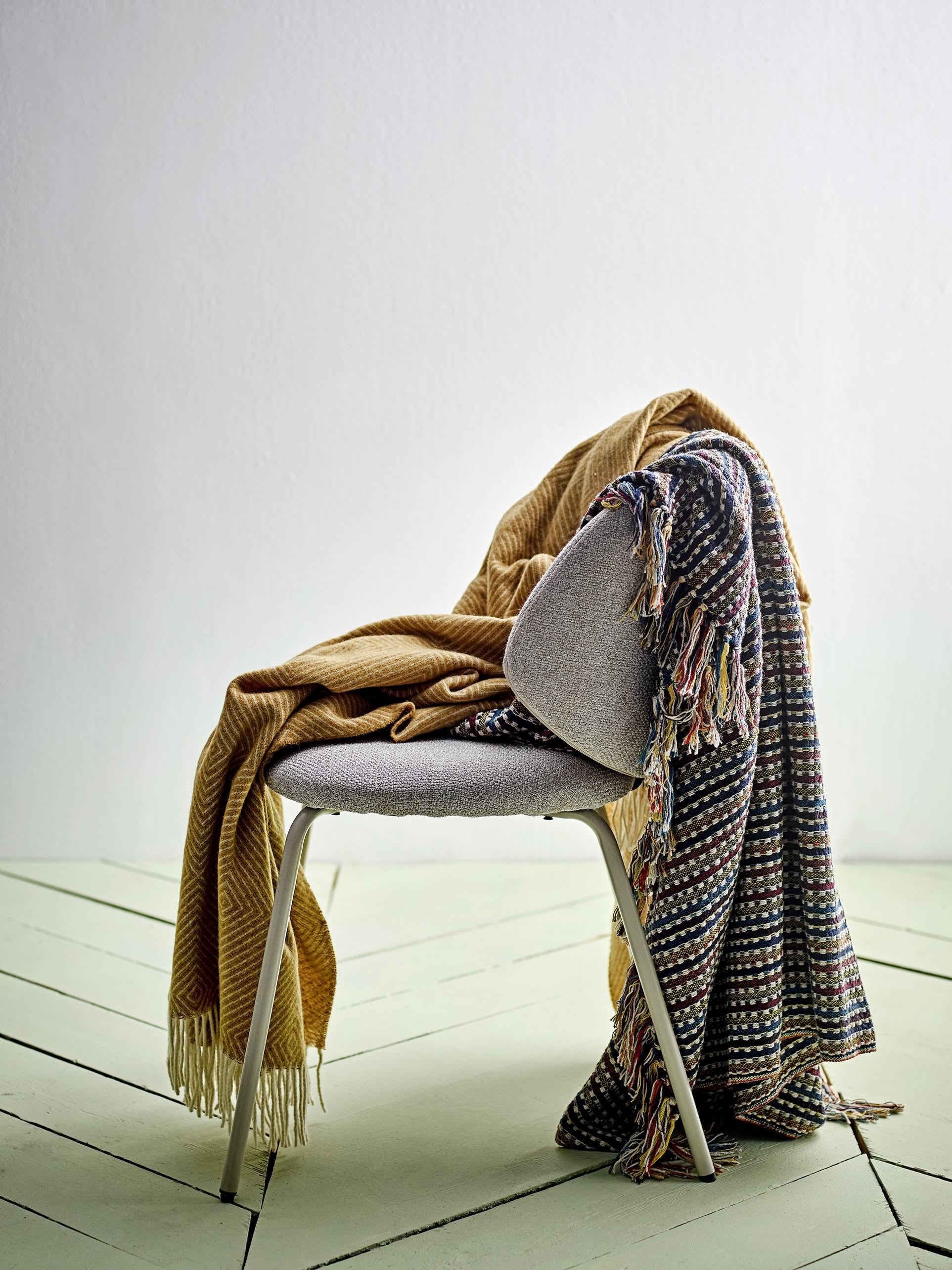A modern gray chair with metal legs is draped with the Bloomingville Lazzario Throw by Bloomingville in multicolored recycled cotton and a mustard yellow fringed blanket, set against a light wall and pale wooden floor.