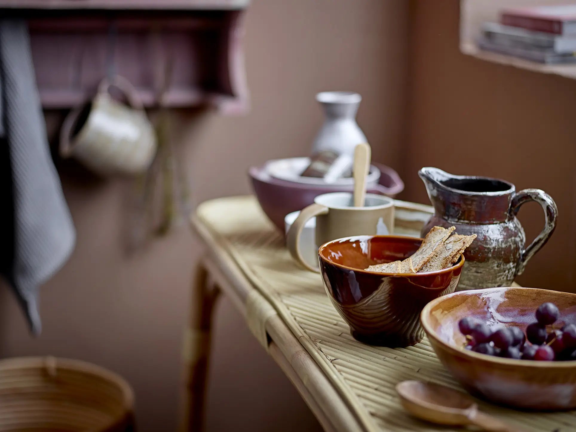 A rustic table set with the Bloomingville Lotus Bowl, Set of 3 by Bloomingville, plus a mug, jug, grapes, and bread slices. Soft window light highlights the cozy kitchen—perfect for serving Asian dishes.