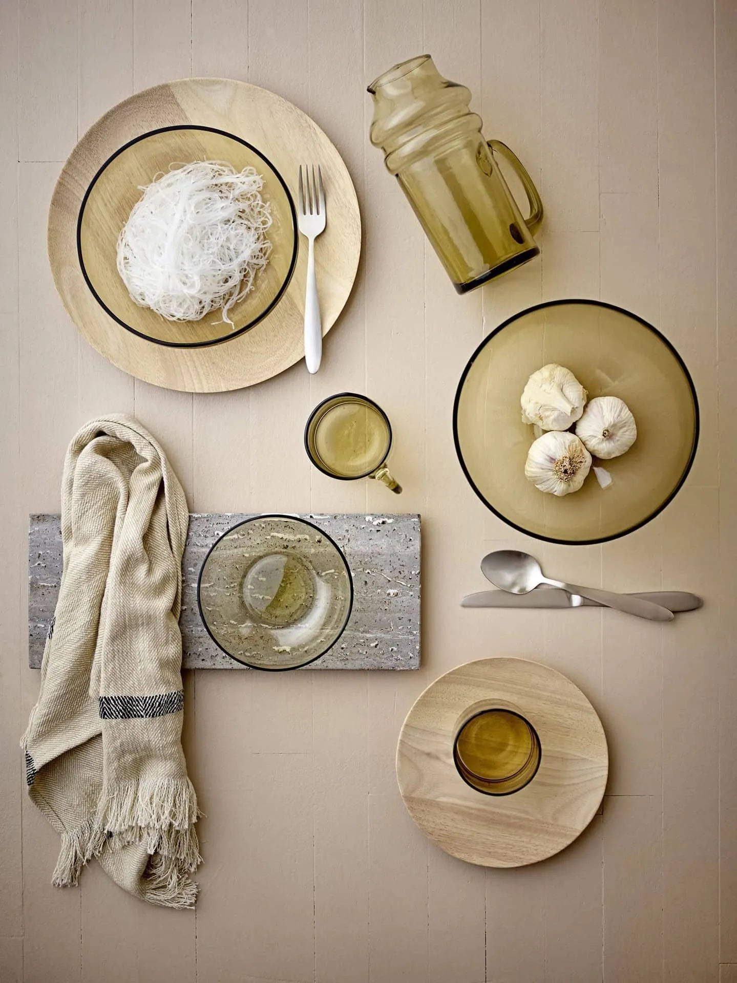 A flat lay shows rice noodles and garlic on a 20cm Bloomingville Nolia Plate with a rustic finish, surrounded by amber glasses, a pitcher, cutlery, and a beige cloth napkin draped over a stone slab.