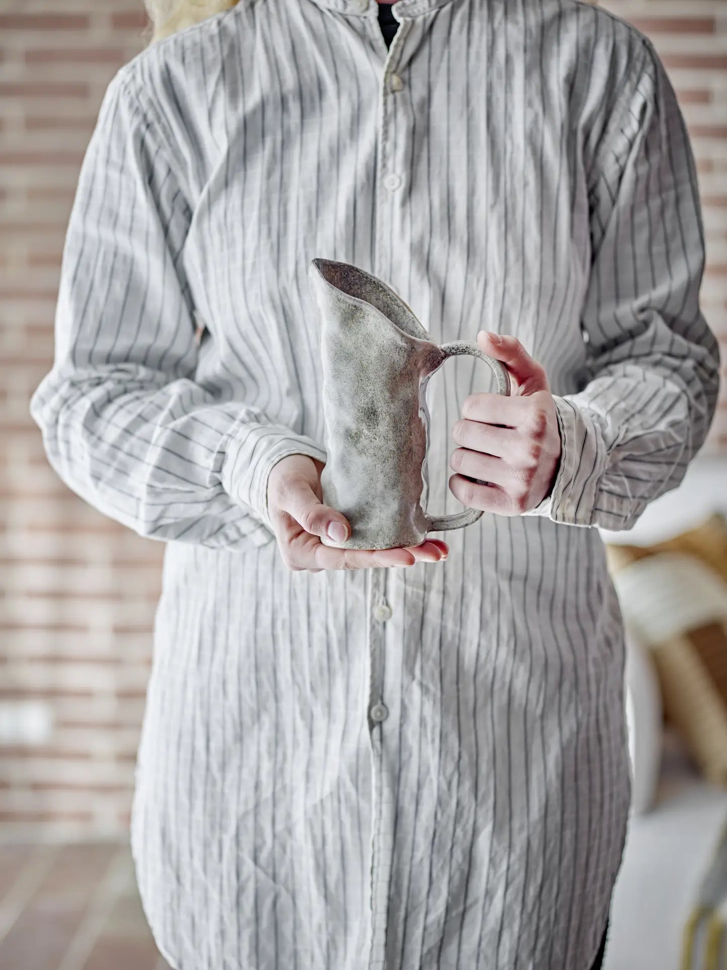 A person in a light-striped button-up shirt holds the Bloomingville Razan Jug from Bloomingville with both hands, set against a backdrop of a brick wall and blurred indoor elements.