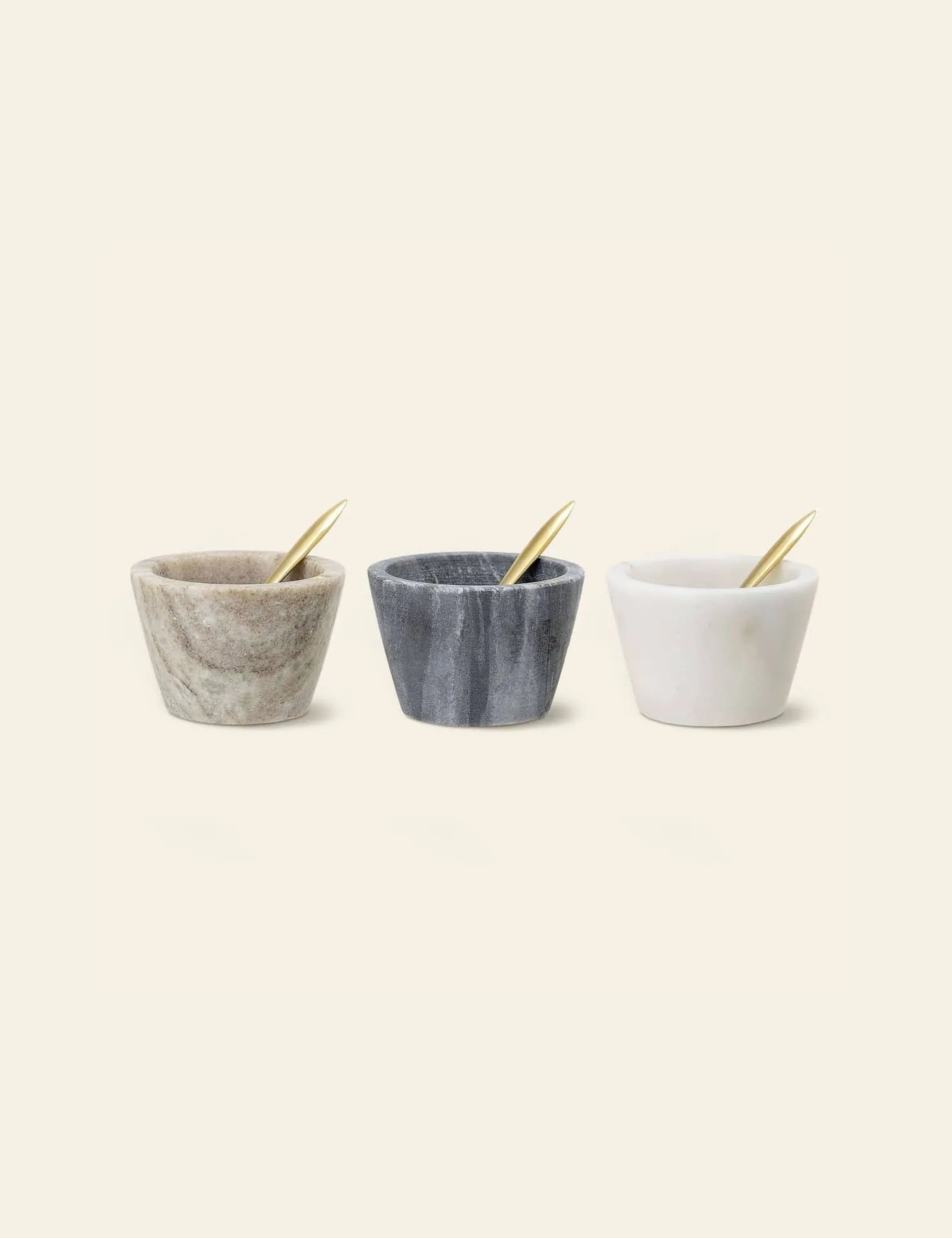 Three Bloomingville Rosamynthe Salt Jars with gold spoons—beige, dark gray, and white—are arranged side by side on a plain background, ideal for serving spices.