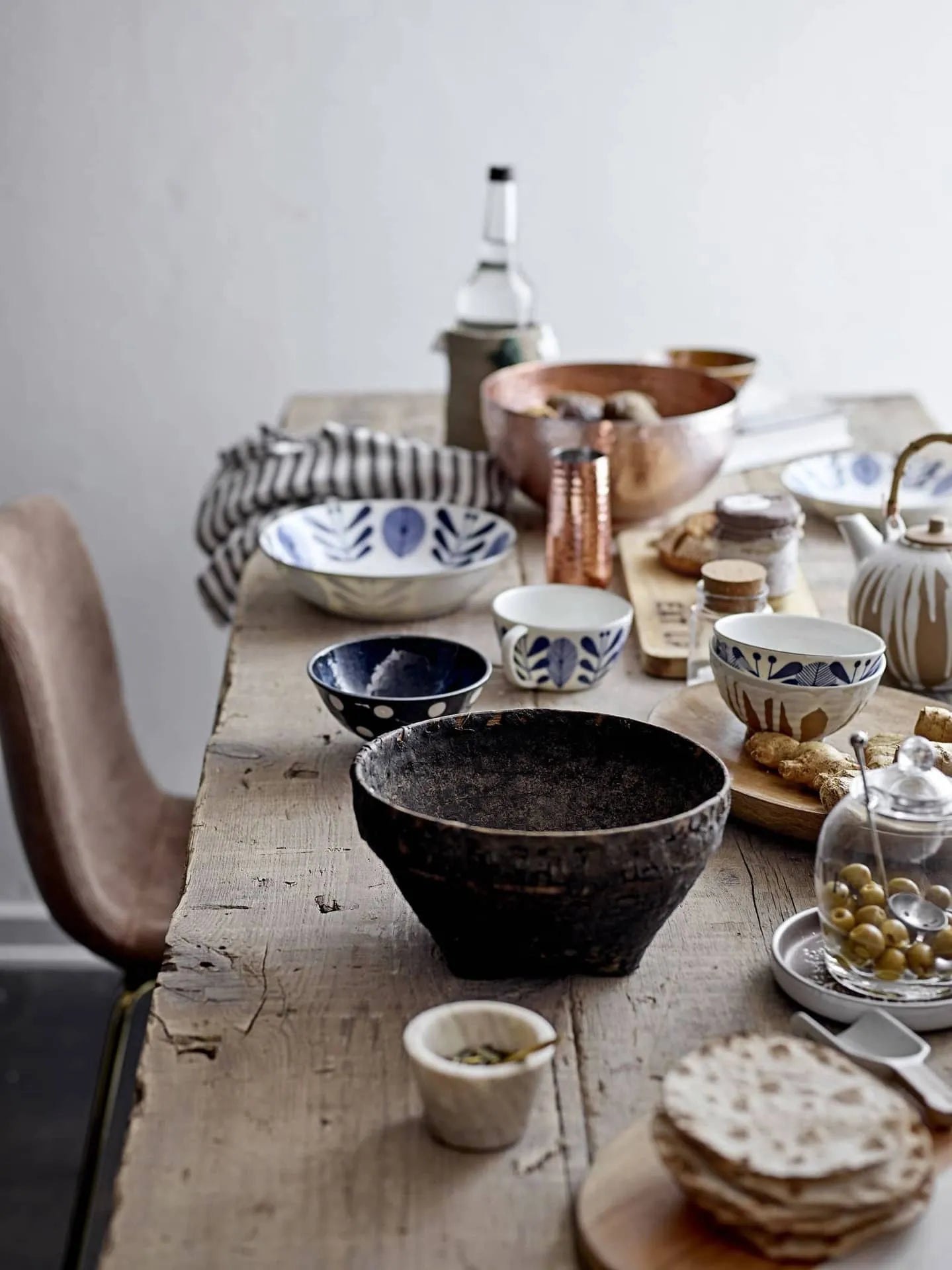 A rustic wooden table features ceramic bowls, a Bloomingville teapot, the Bloomingville Rosamynthe Salt Jar w/Spoon, glass jars, spices, crackers, olives, and ginger. A striped cloth, copper pot, and chair complete the cozy dining setting.