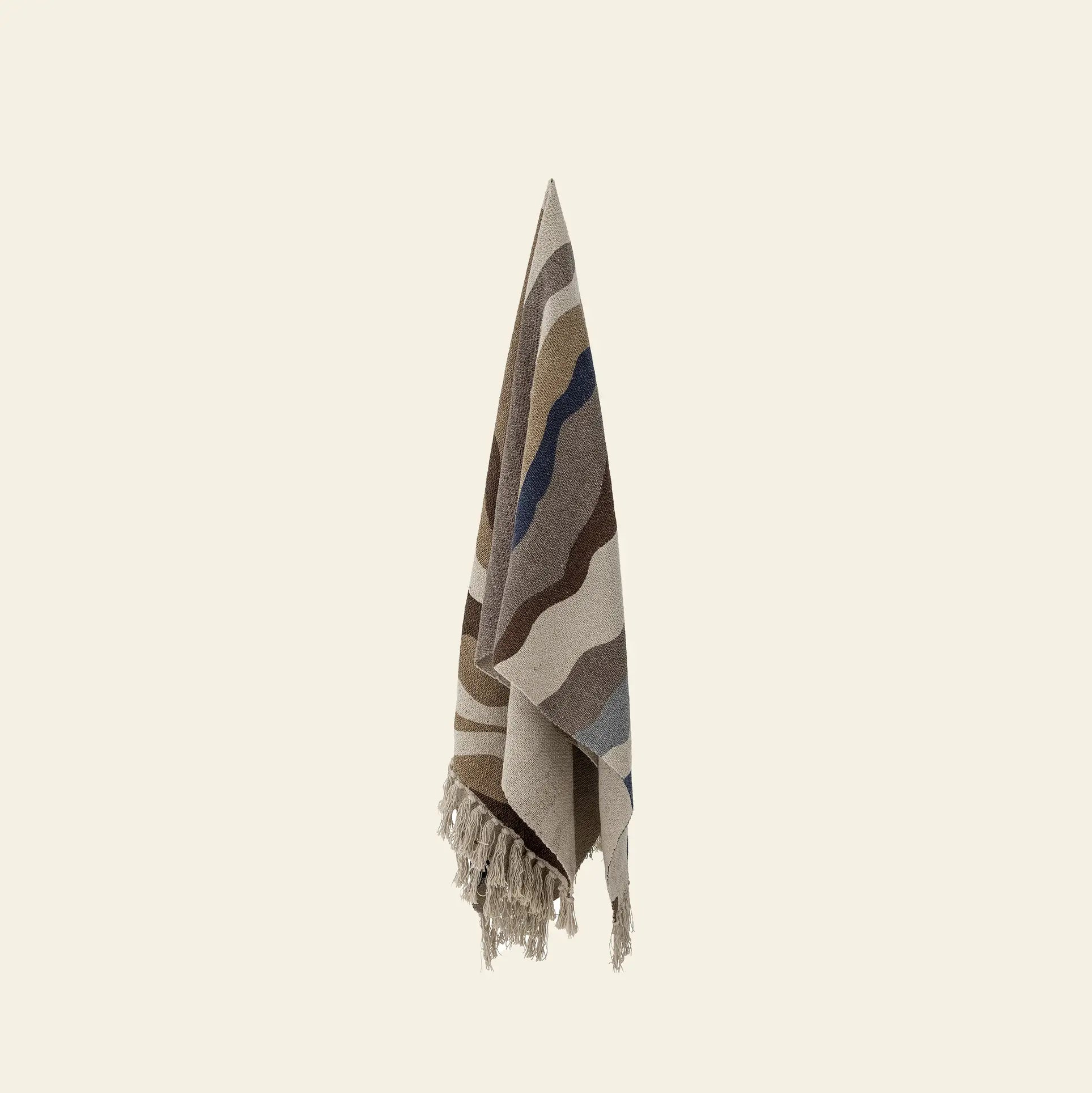 The Bloomingville Stephania Throw by Bloomingville features beige, brown, and blue wavy stripes on recycled cotton, with fringed edges. It hangs vertically against an off-white background.