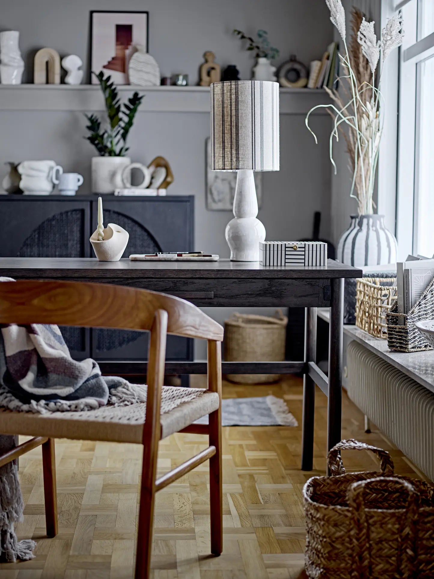 A cozy home office with a wooden desk, chair, books, baskets, plants, decor—and a Bloomingville Stephania Throw in recycled cotton with a printed pattern—brings warmth to the gray wall lit by natural light.