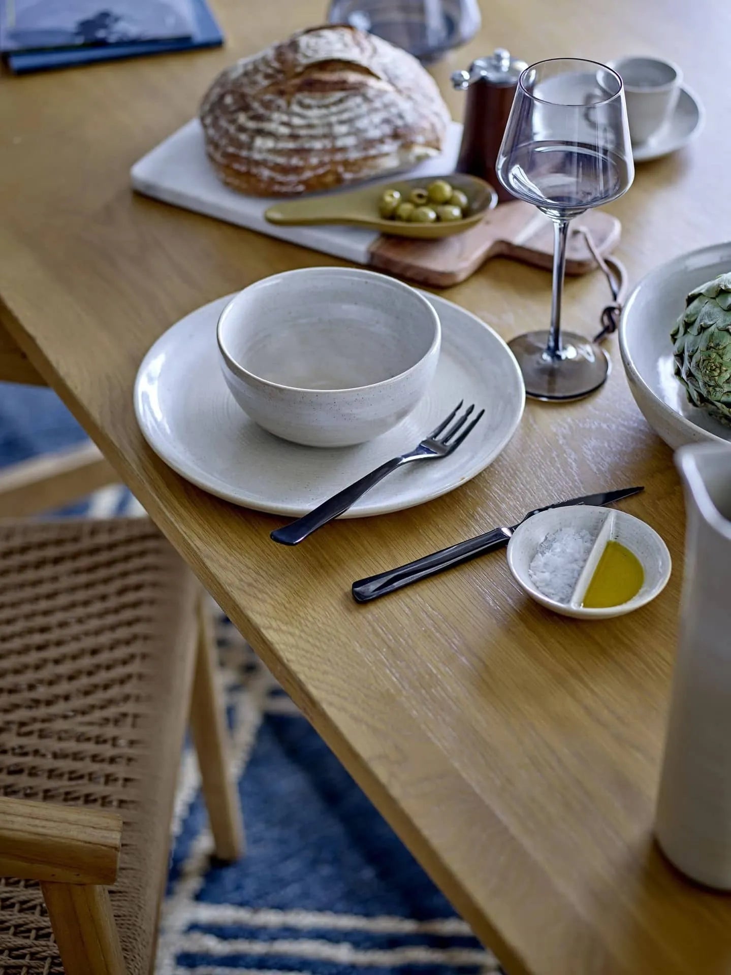 A wooden dining table is set with a Bloomingville Taupe Plate, matching bowl, fork, knife, wine glass, bread, olives, artichoke, and olive oil dish. A woven chair and blue patterned rug complete the charming setting.