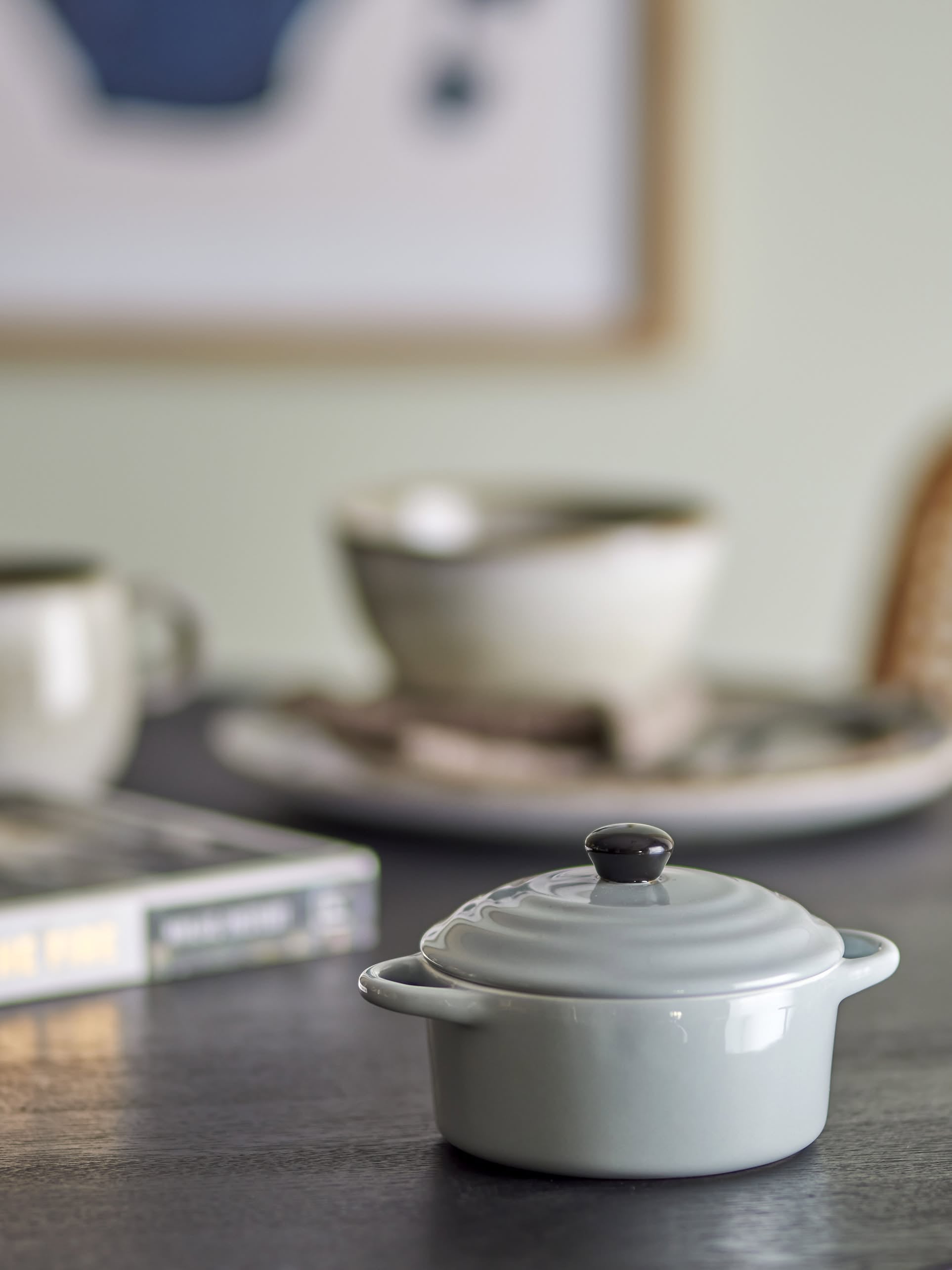 The Bloomingville Thanos Oven Dish w/Lid, a small gray stoneware pot, sits on a table with a blurred mug, bowl, plate, and book in the background—creating a cozy setting perfect for casseroles or oven dishes.