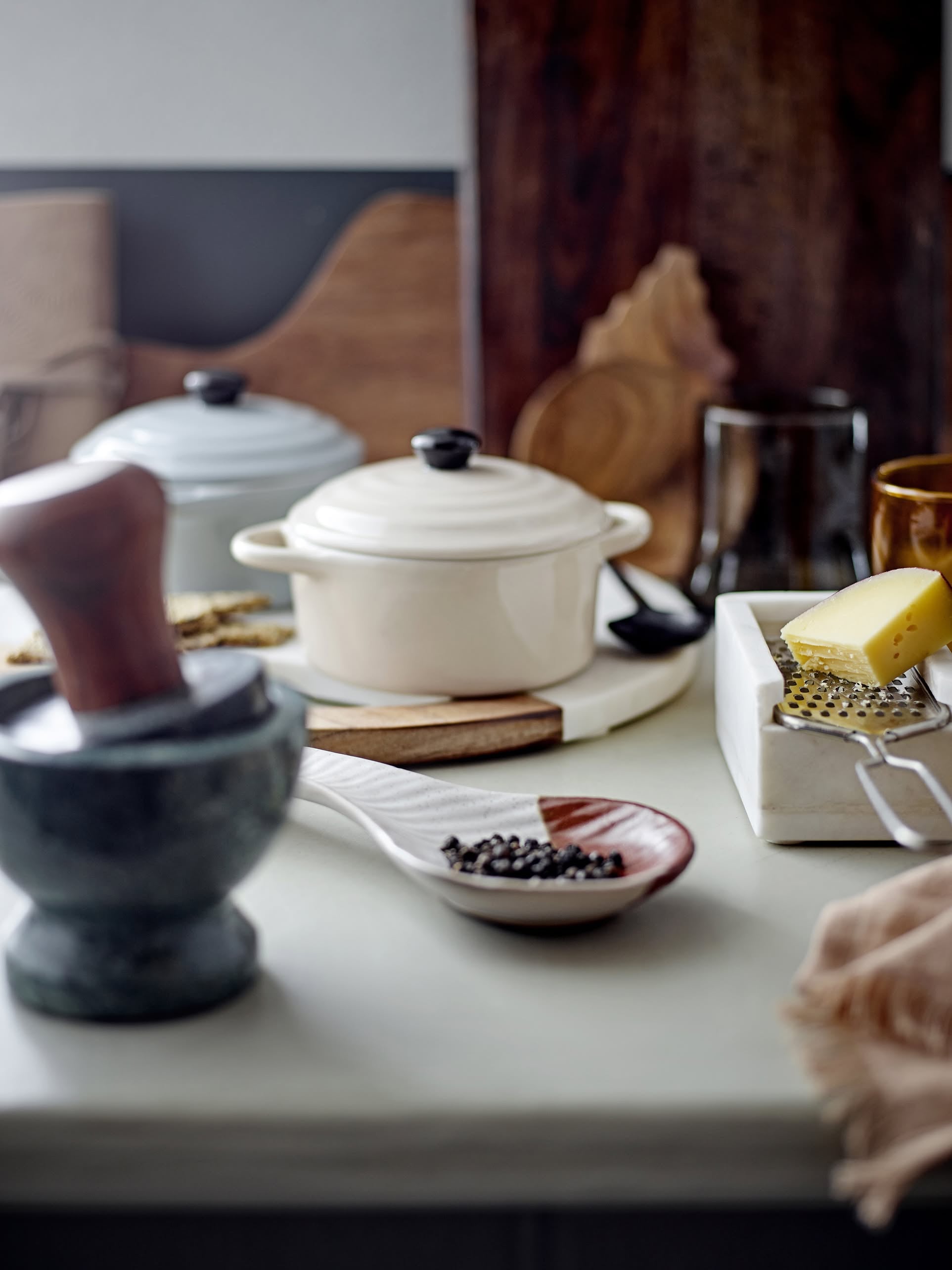 A kitchen countertop with a marble mortar and pestle, a white spoon with black peppercorns, a butter dish and grater, and Bloomingville Thanos Oven Dishes with Lids by Bloomingville displayed elegantly in the background.