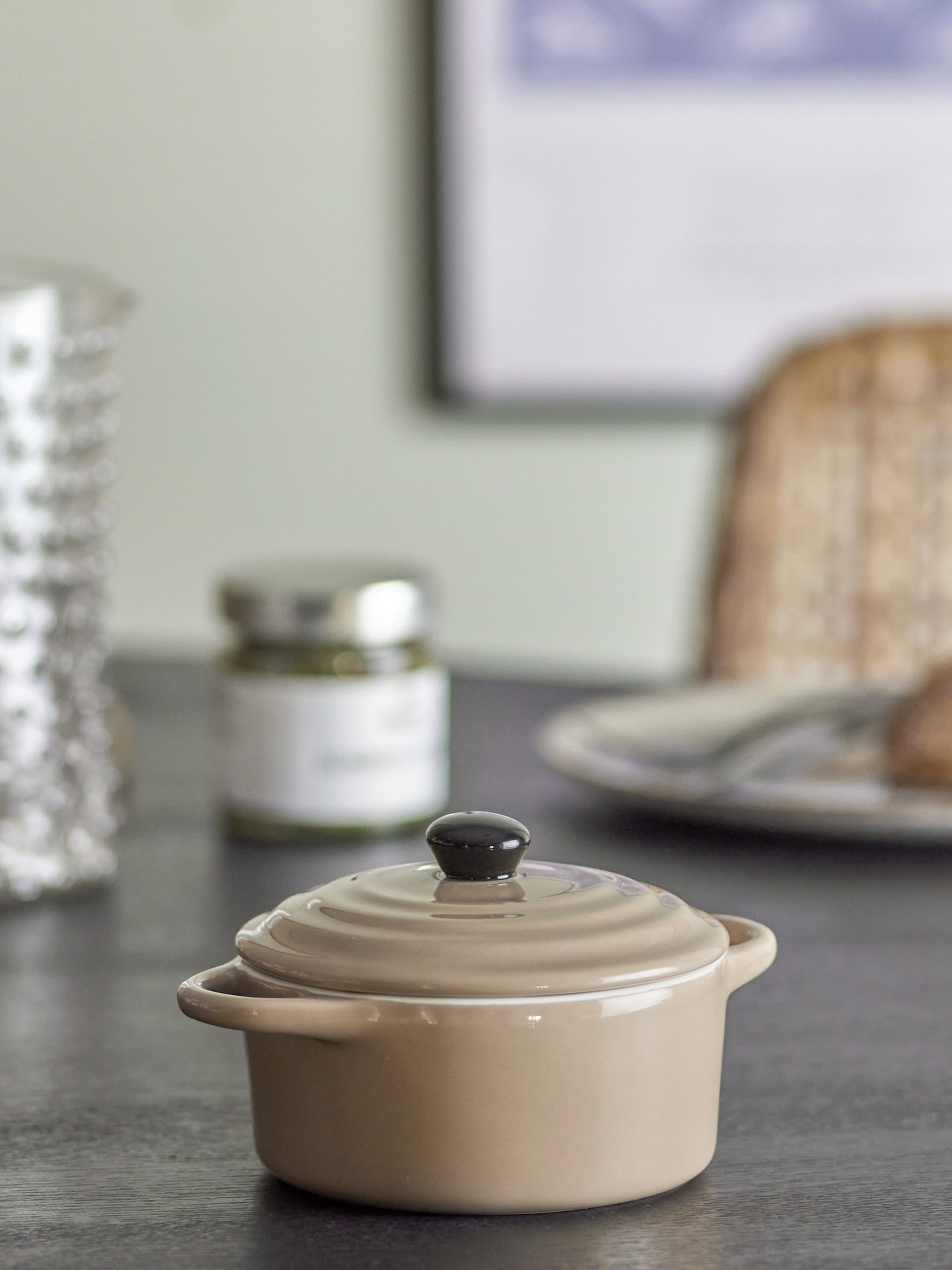 The Bloomingville Thanos Oven Dish w/Lid, a small beige stoneware pot, sits on a dark countertop. In the blurred background, a glass jar and plate of bread create a cozy kitchen scene perfect for casseroles.