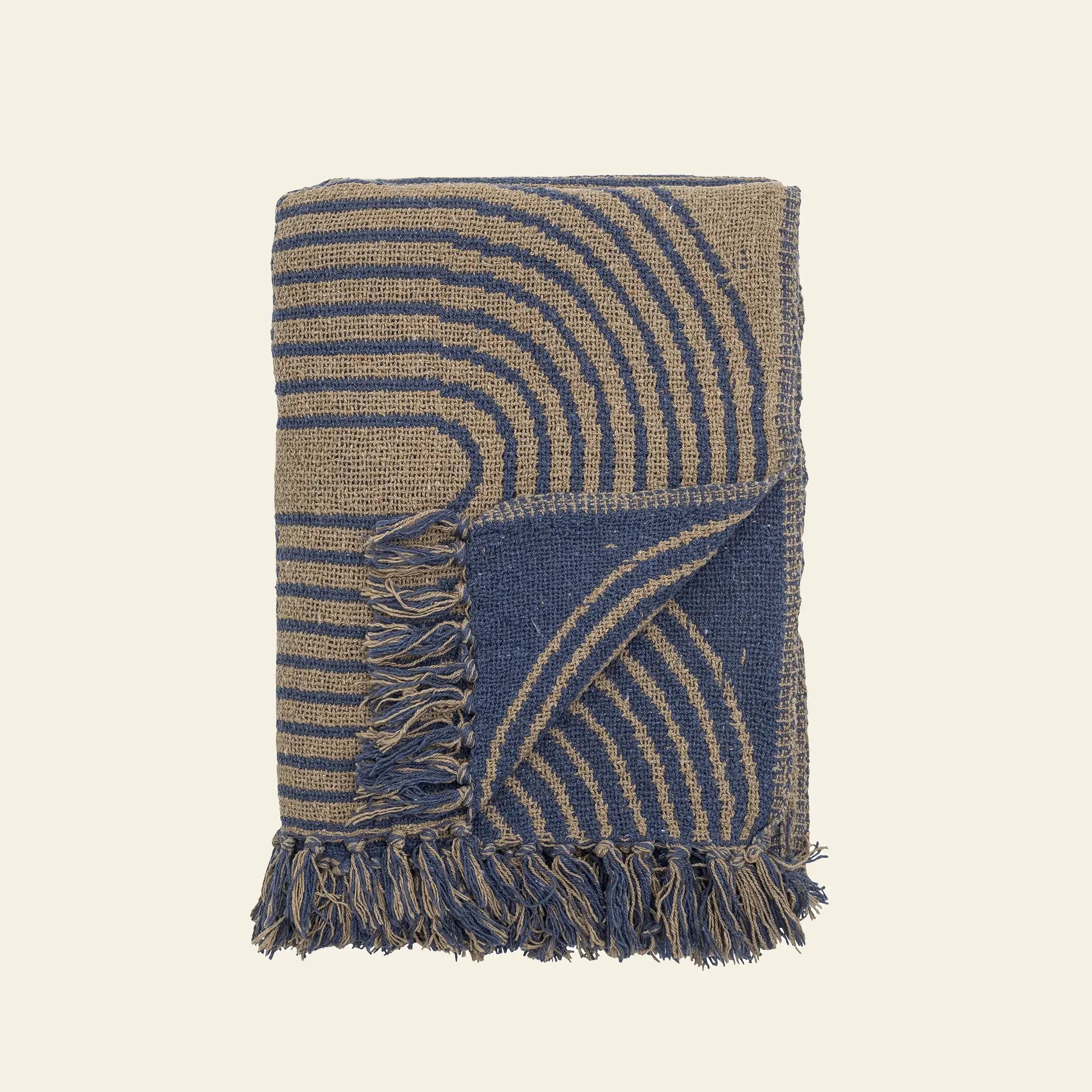 The Bloomingville Tiffanie Throw by Bloomingville is a cotton woven blanket with blue and beige curved stripes in a graphic pattern, fringed edges, and a turned-up corner showing the reverse side, displayed against a plain light background.