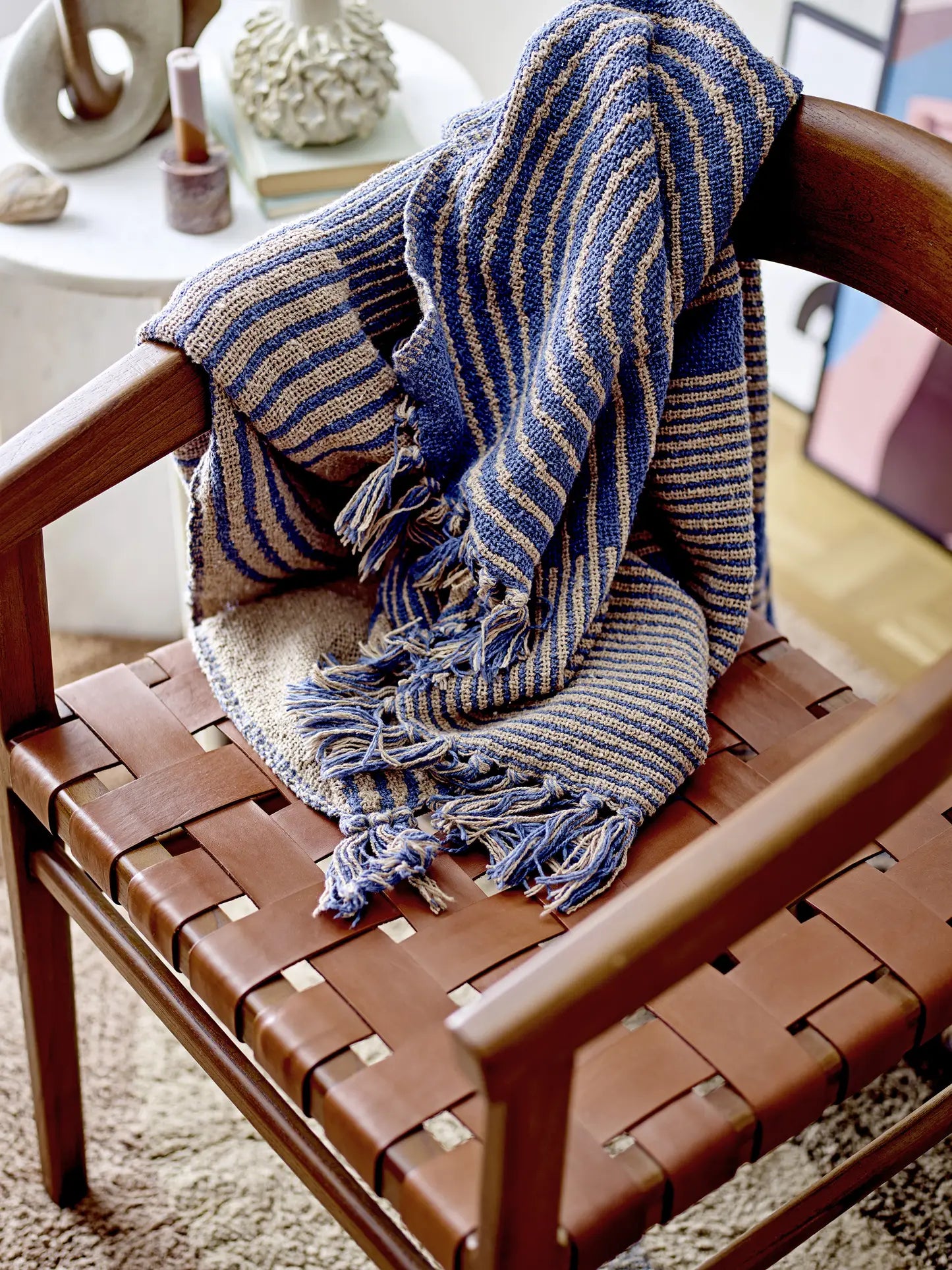 The Bloomingville Tiffanie Throw by Bloomingville, featuring blue and beige stripes with fringed edges, is draped over a wooden chair with a brown woven leather seat, set on a textured beige rug next to a small round table.