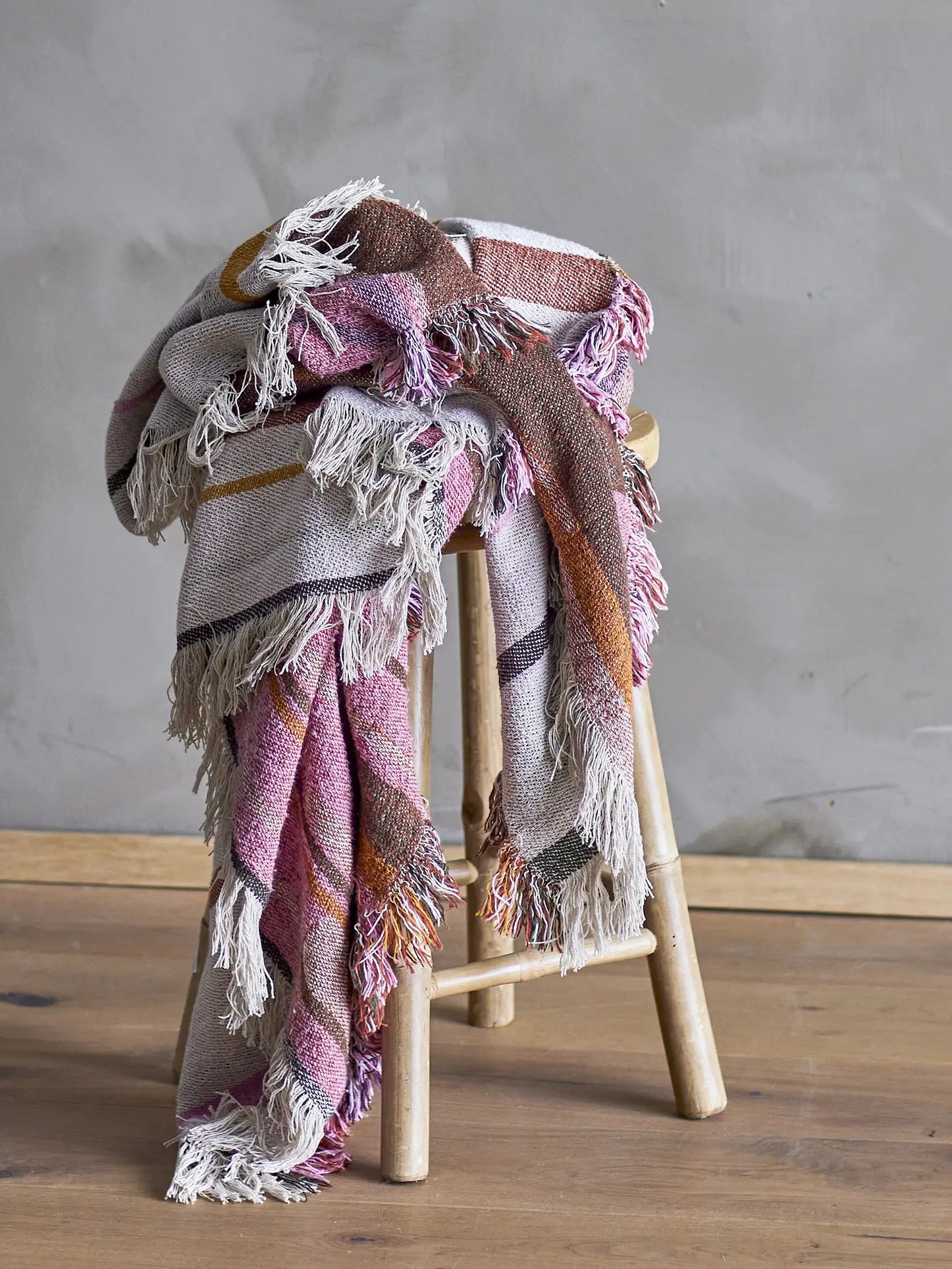 The Bloomingville Toscana Throw, a multicolored woven blanket made from recycled cotton with fringed edges, is displayed over a wooden stool against a gray textured wall and wooden floor.