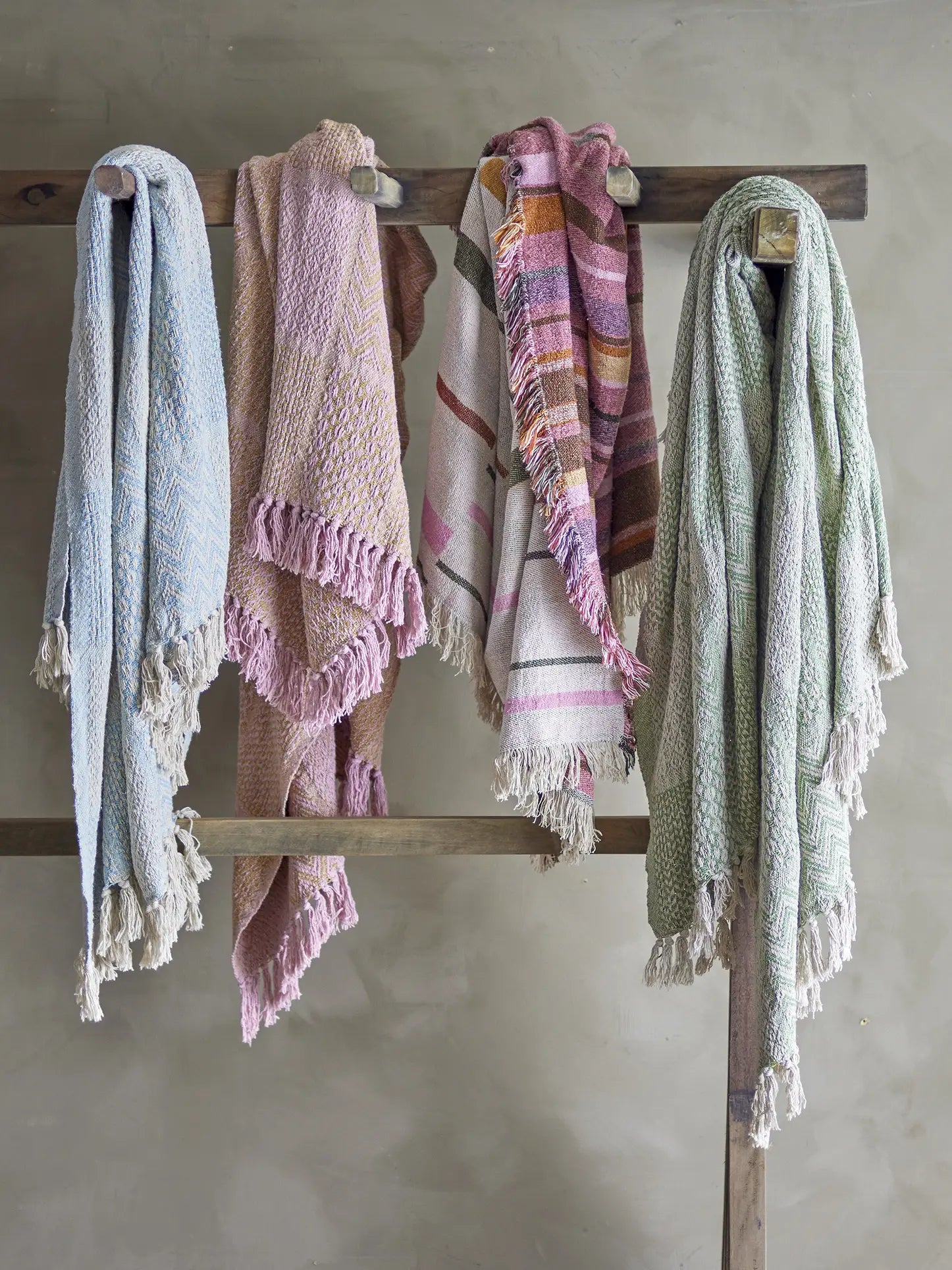 Four Bloomingville Toscana Throws—woven from recycled cotton in pastel blue, pink, multicolor stripes, and green—hang neatly over a rustic rack against a neutral wall.