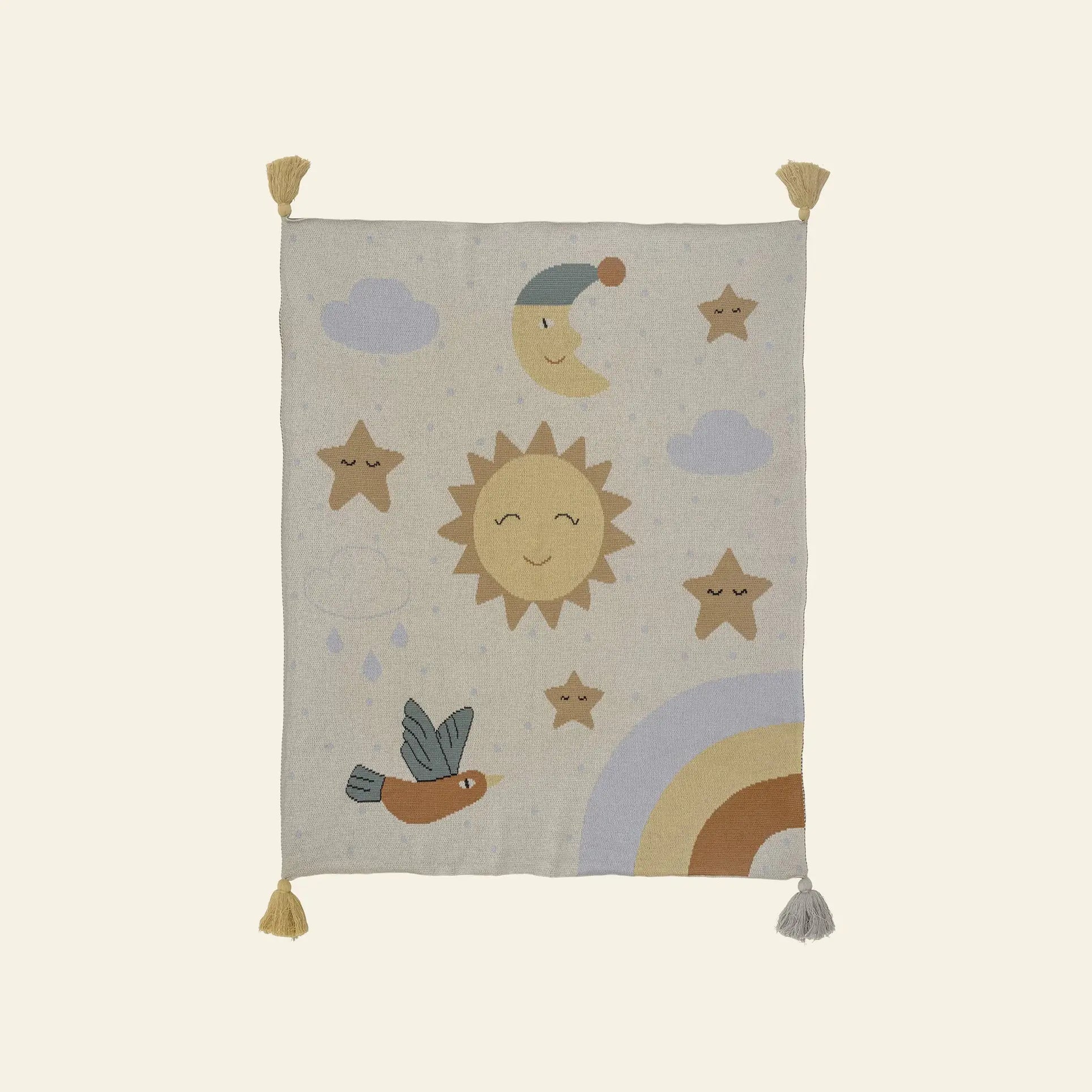 The Bloomingville Vigge Throw by Bloomingville is a beige OEKO-TEX certified cotton blanket with corner tassels, featuring pastel sky-themed designs like a smiling sun, moon in nightcap, stars, clouds, bird, raindrops, and rainbow.